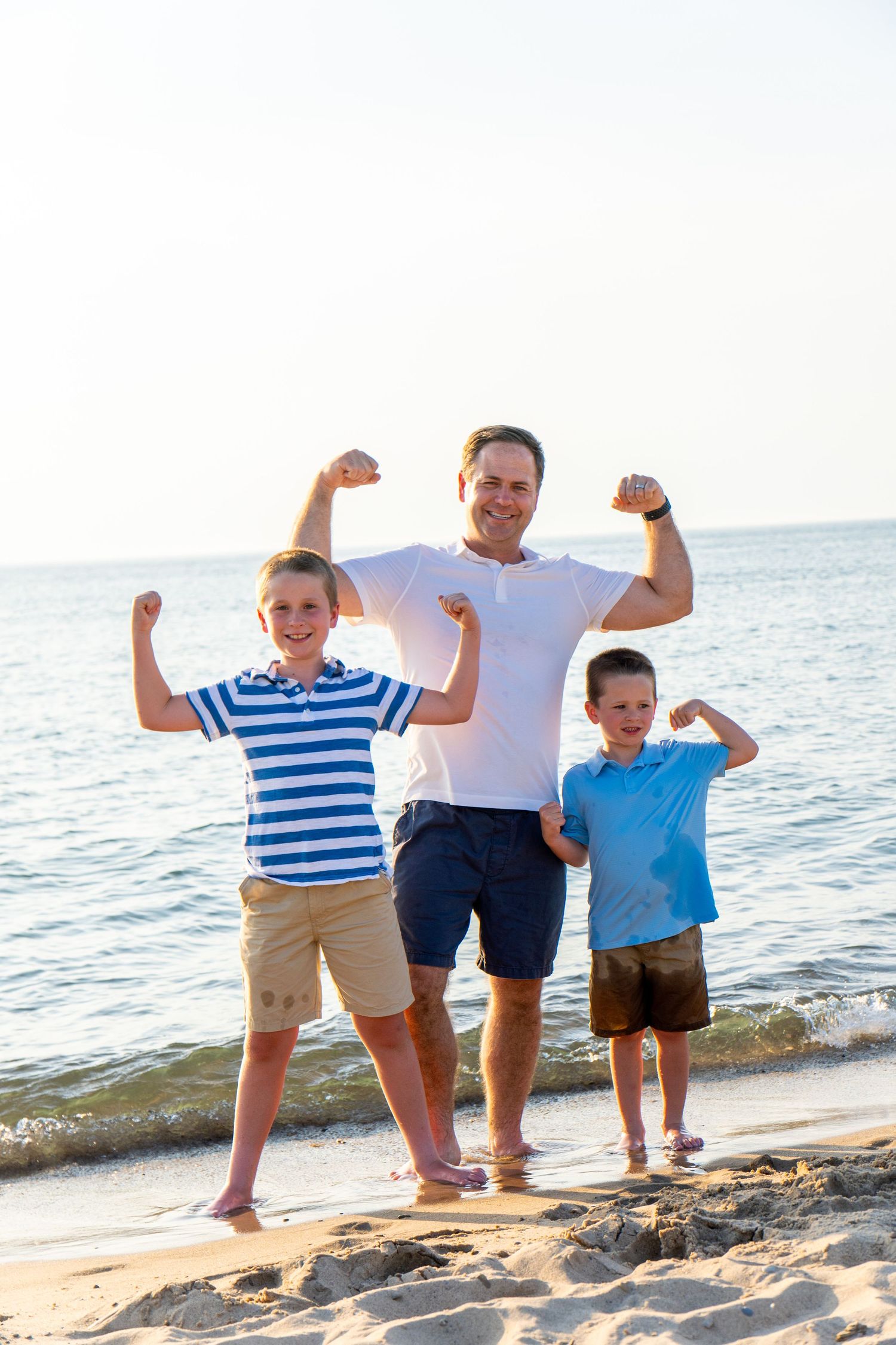 A smiling man and two young boys flex their arms in a muscle pose while standing barefoot on a sandy beach at sunset.