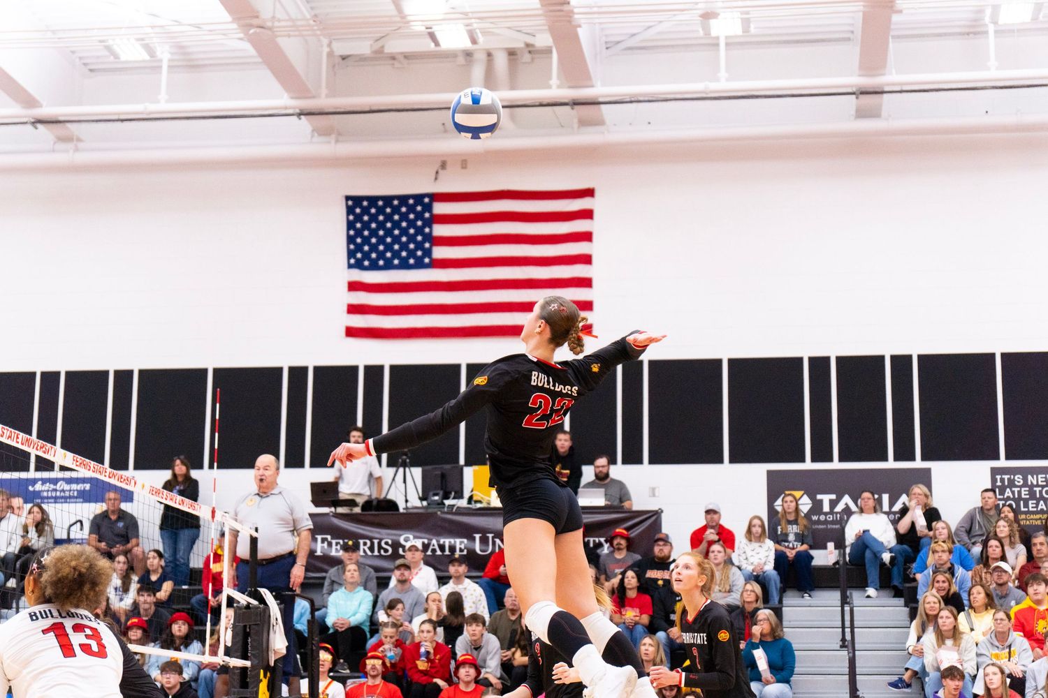 Volleyball player in a black jersey leaps to spike the ball during an indoor college match as spectators and an American flag fill the gym.