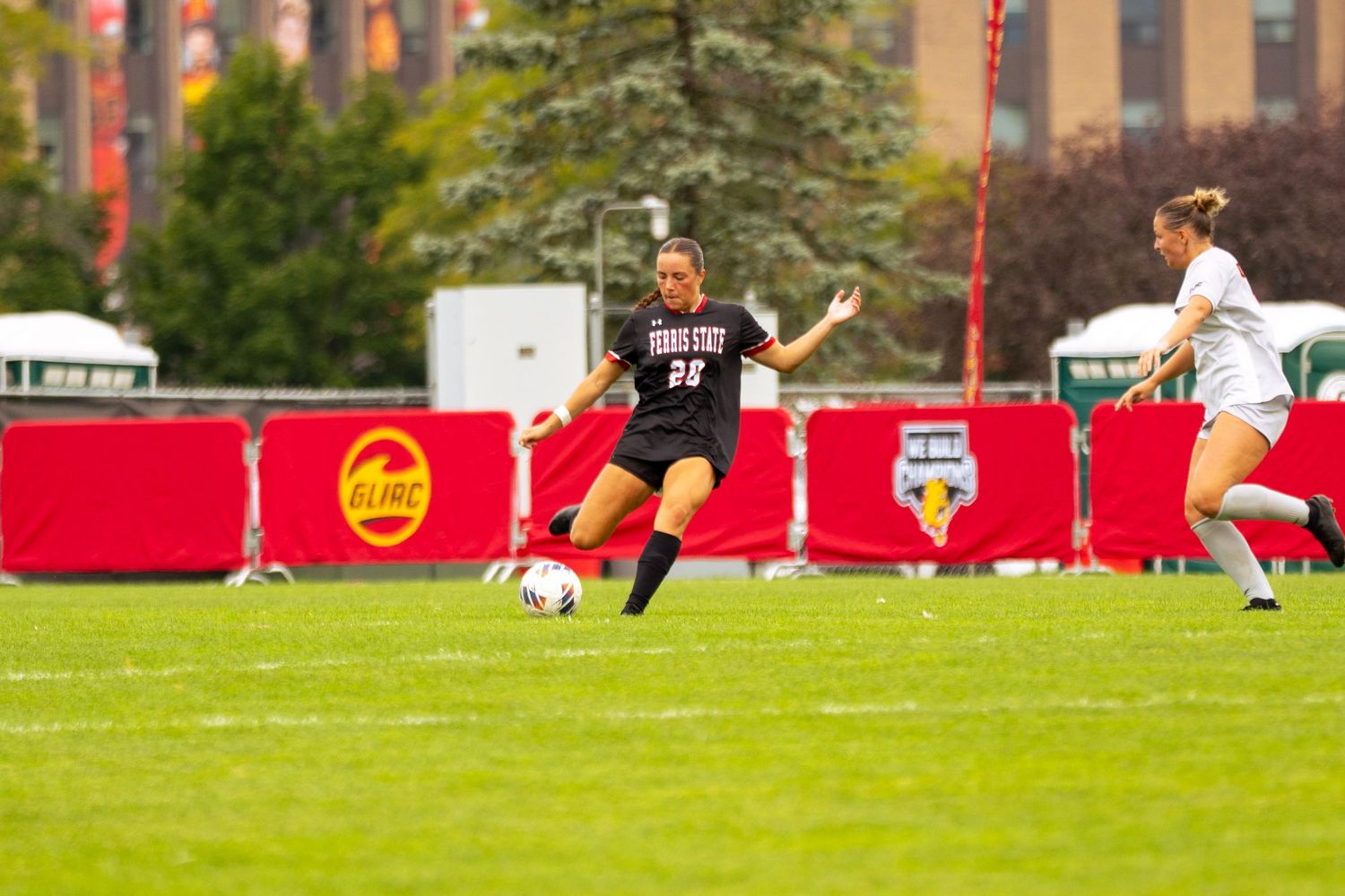 Female soccer player in a dark uniform kicks the ball on a green field while an opponent in white looks on during a college match.