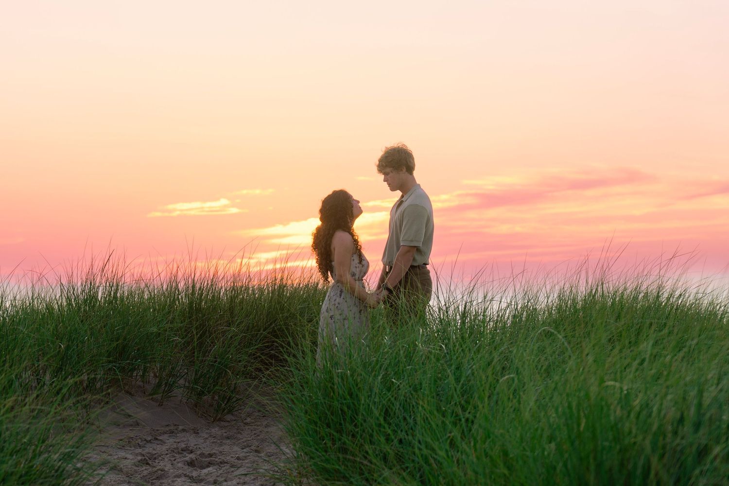 Two silhouetted figures standing in a field of tall grass against a vibrant sunset sky during golden hour.