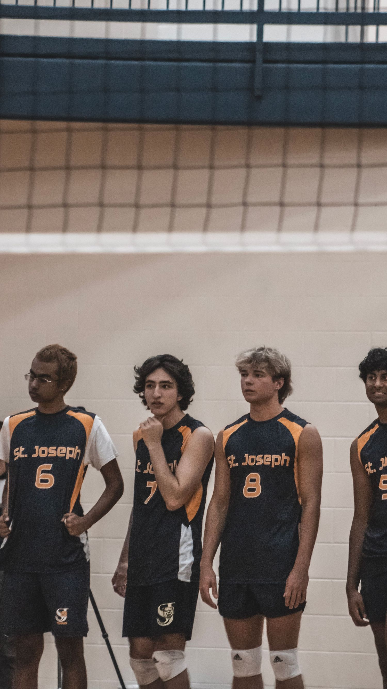 Four volleyball players in black St. Joseph jerseys line up against wall during match.