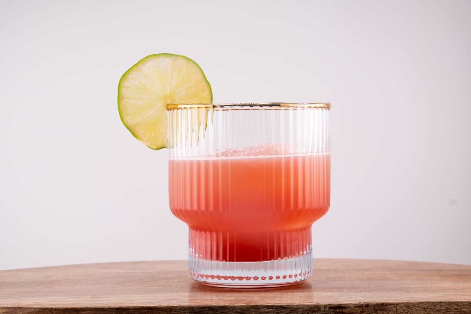 A pink cocktail garnished with a lime wheel in a ribbed glass tumbler.