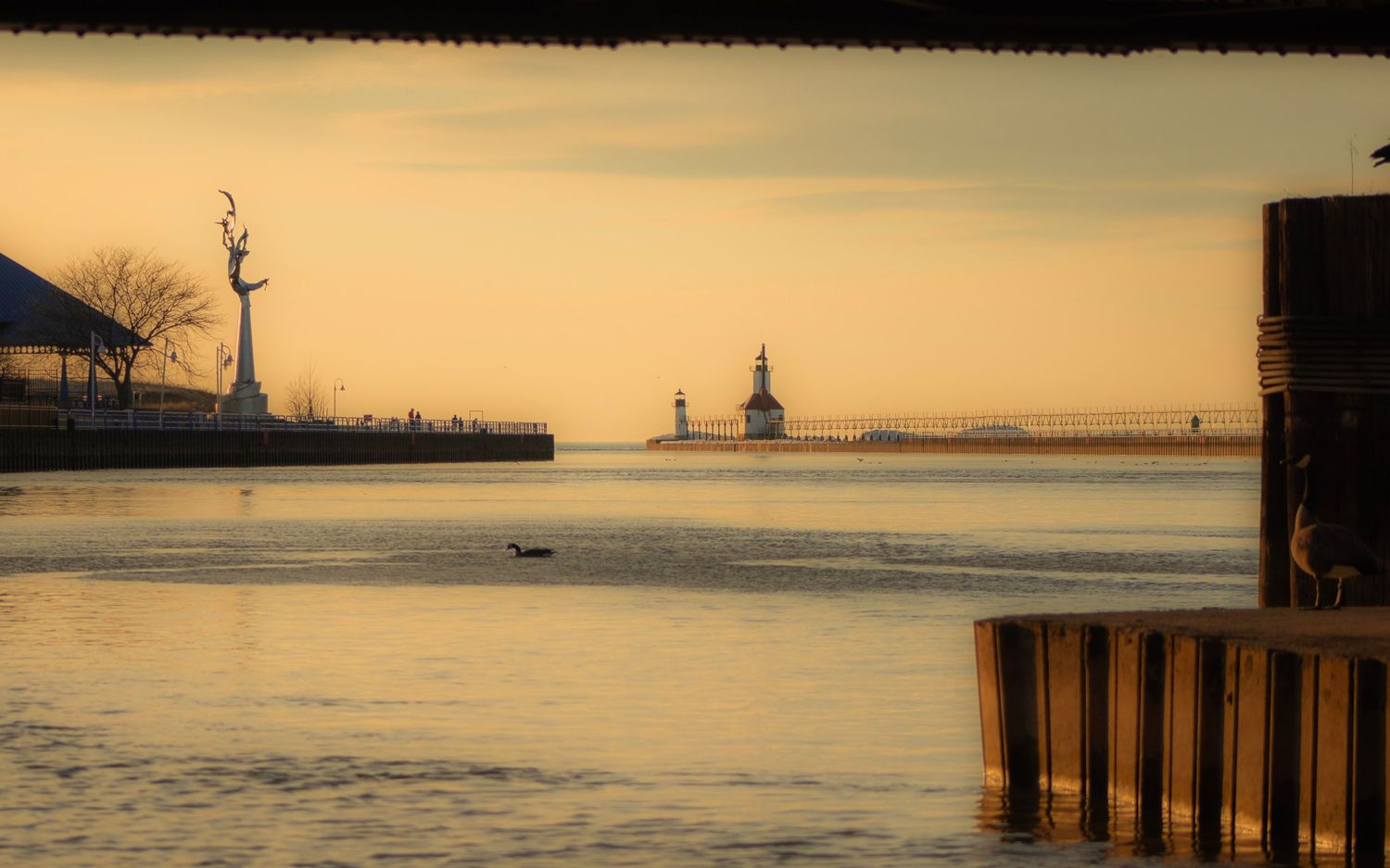 Harbor lighthouse silhouetted against golden sunset sky with wooden pilings in foreground.