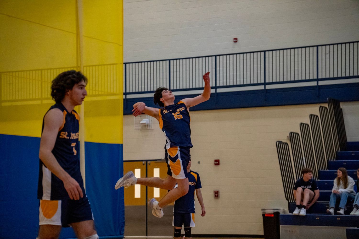 Two volleyball players leap for the ball during an indoor match with spectators watching in the background.