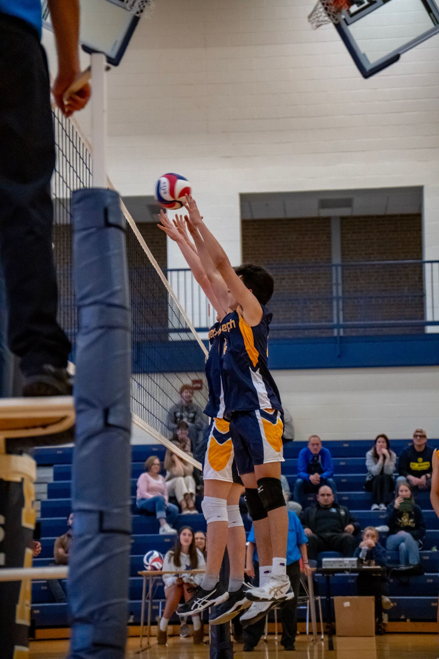 Two players leap up to block a volleyball at the net during an indoor match.