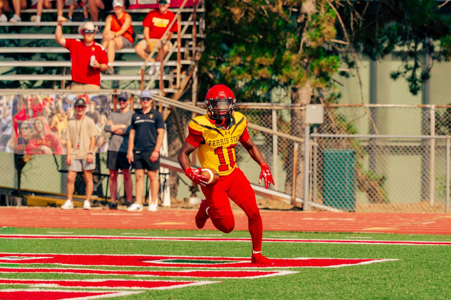 Football player in red uniform runs with ball during game on sunny field with spectators watching from sidelines.