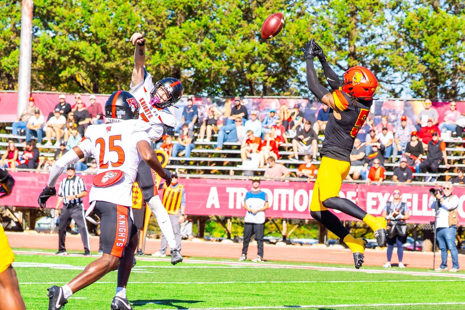 Football players in orange and black leap for a pass during a college game as fans watch from the stands on a sunny day.