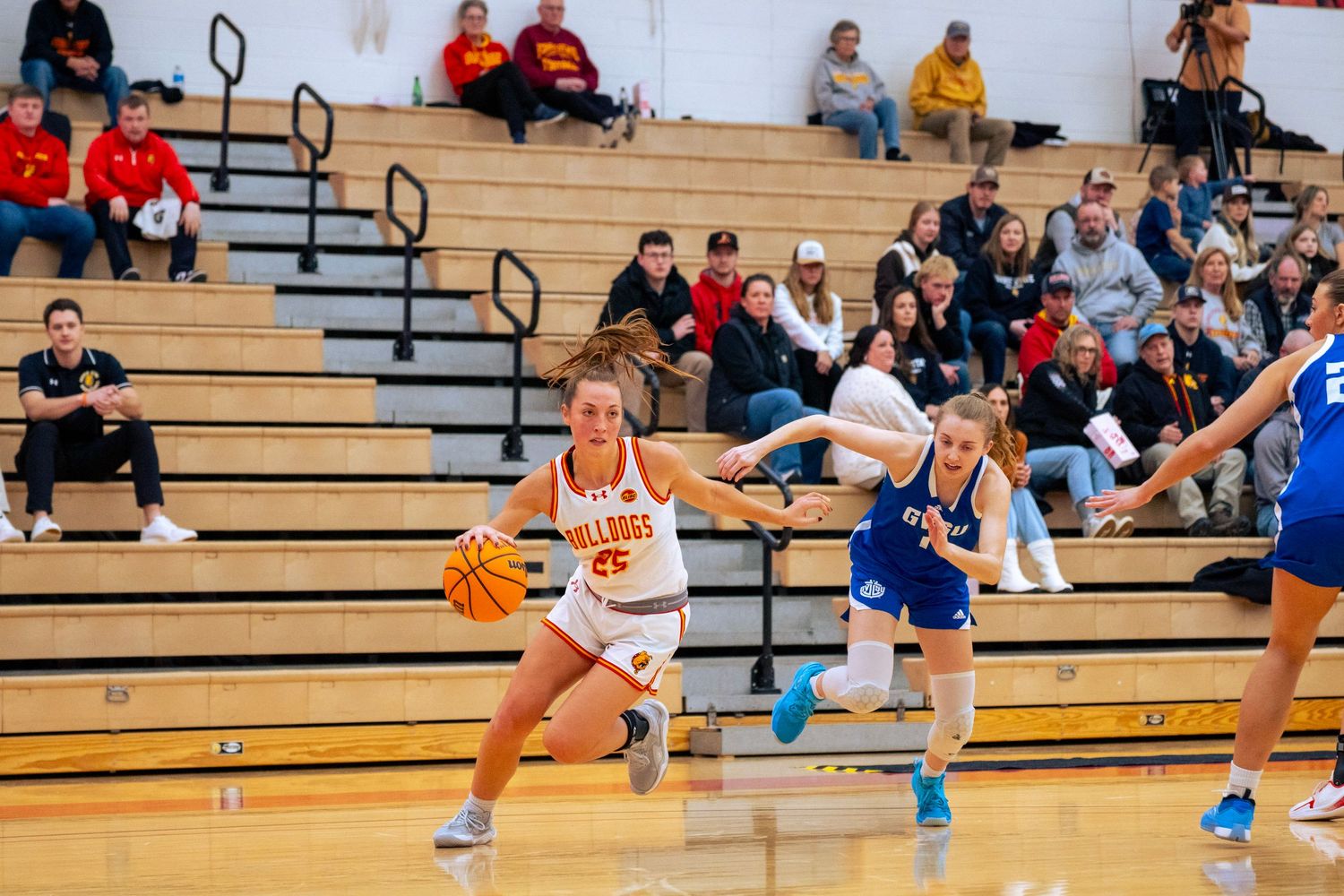 Women's basketball player in a white jersey dribbles past a defender in blue while spectators watch from the gym bleachers.