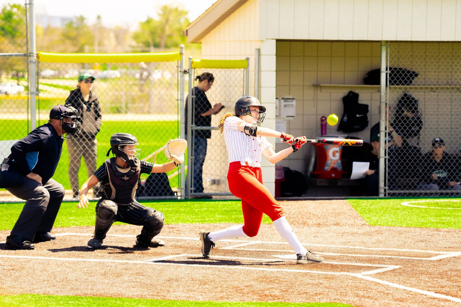 Softball batter in a white jersey and red pants swings at a pitch as the catcher and umpire crouch behind her during a sunny game.