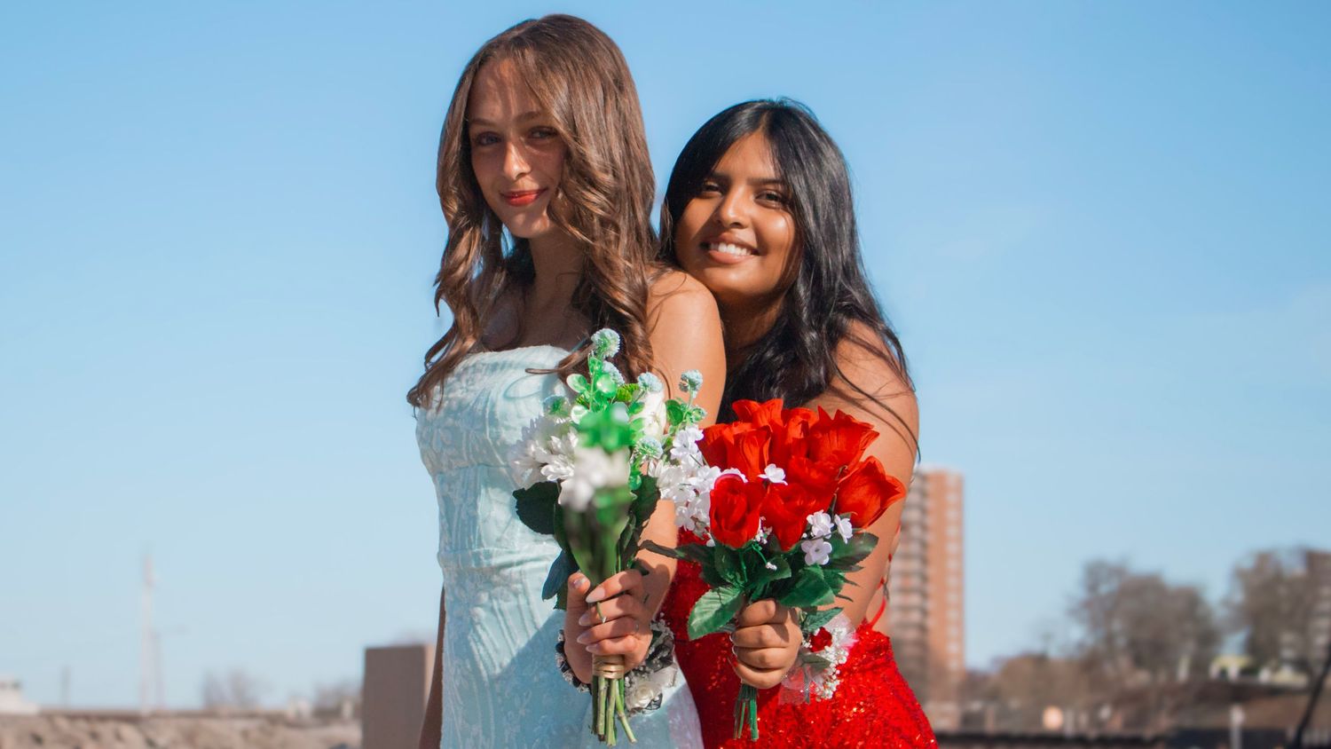 Two friends stand together outdoors against a blue sky holding colorful bouquets of roses and wearing light colored dresses.