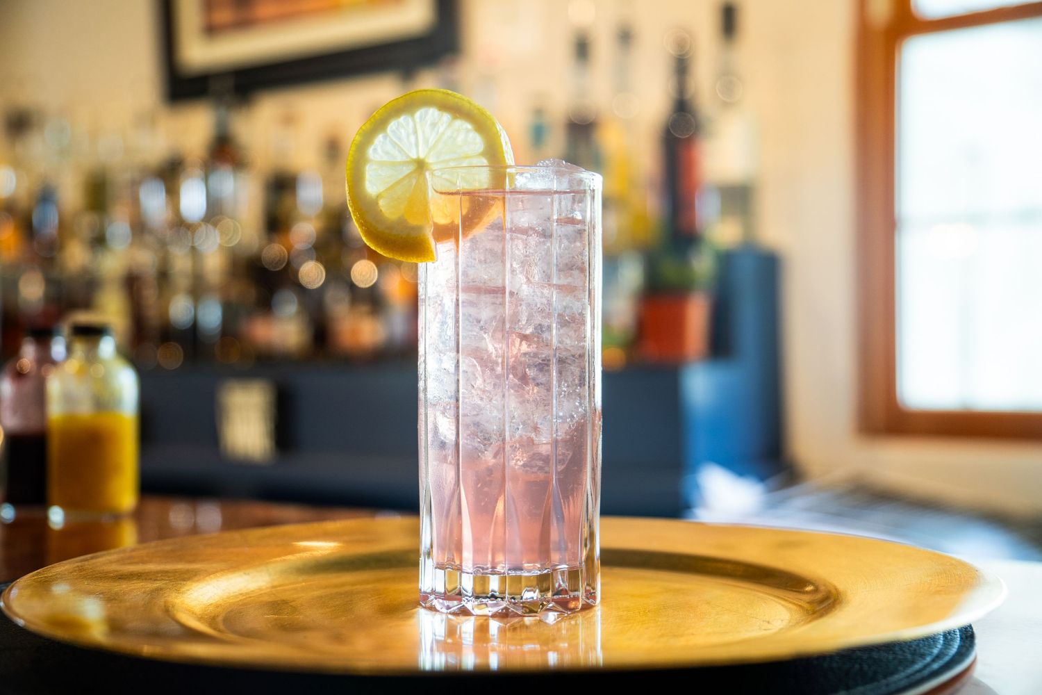 Pink cocktail served on a gold tray with lemon garnish and ice.