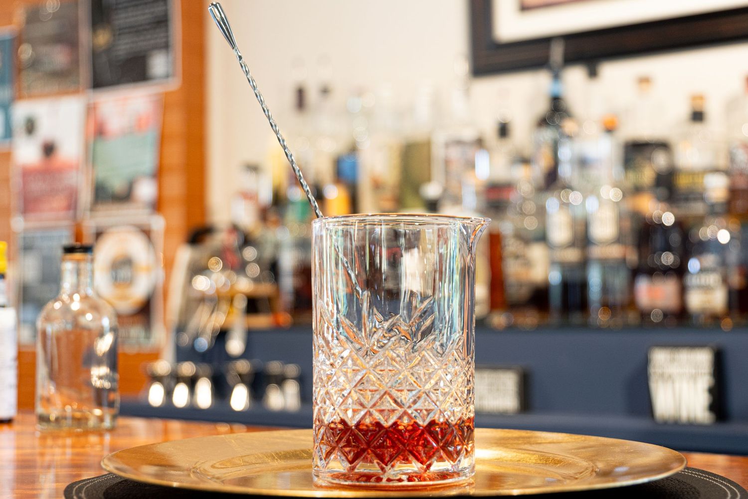 Crystal mixing glass filled with red cocktail syrup on a golden tray in a bar setting.