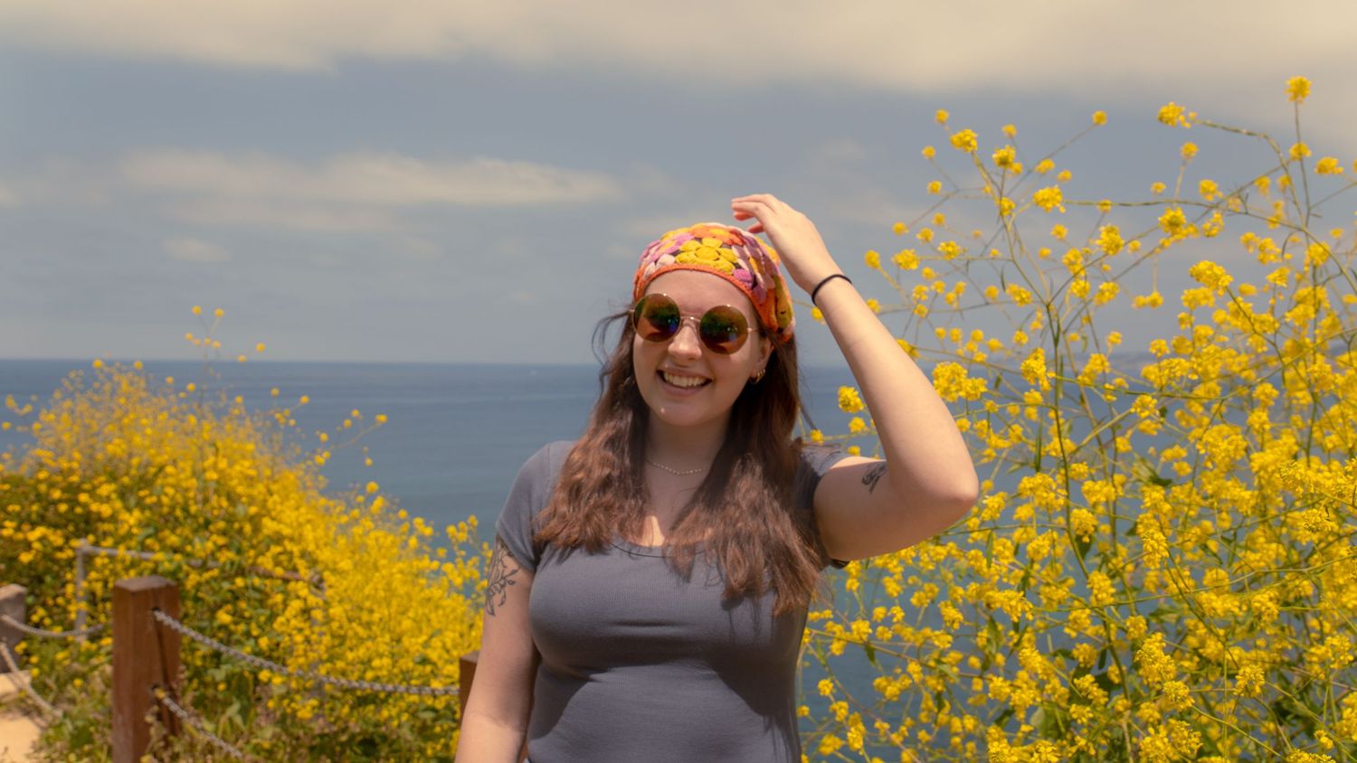 Person in orange headscarf poses joyfully among yellow wildflowers with ocean view.