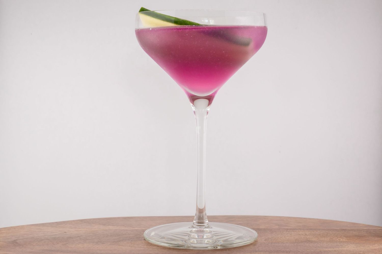 Pink cocktail in coupe glass garnished with cucumber slice on wooden surface.