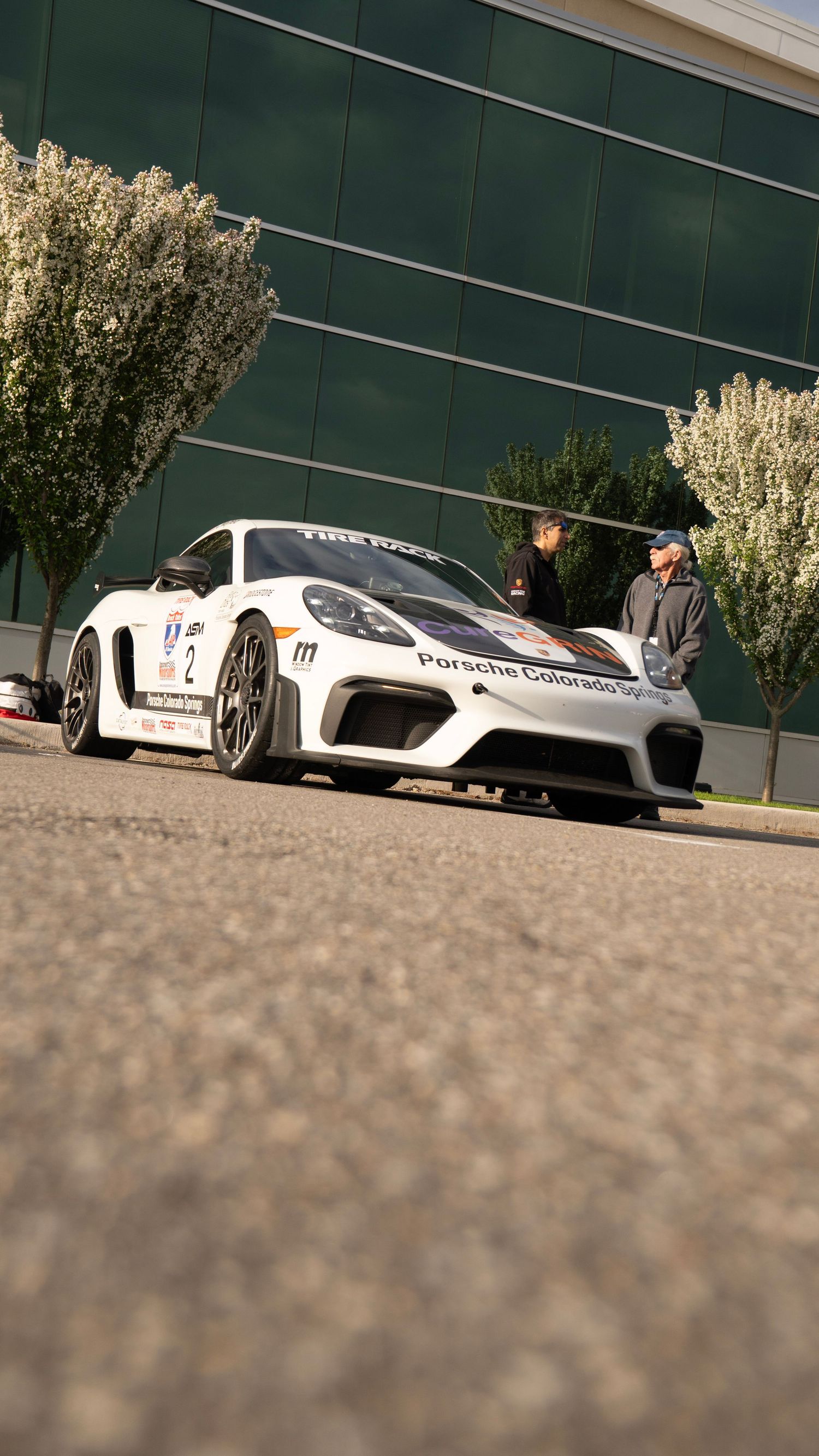 White Porsche GT race car parked in front of a green building with trees.