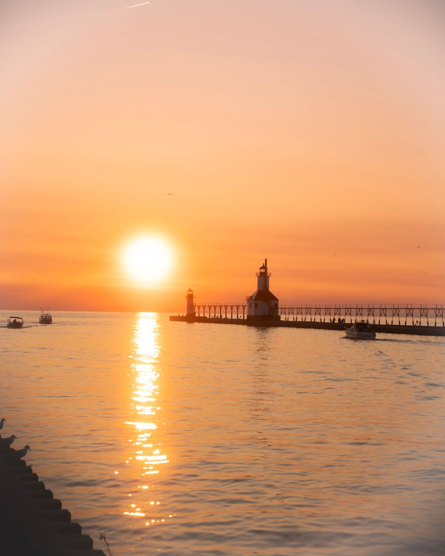 Historic lighthouse silhouetted against brilliant orange sunset with boats on calm waters.