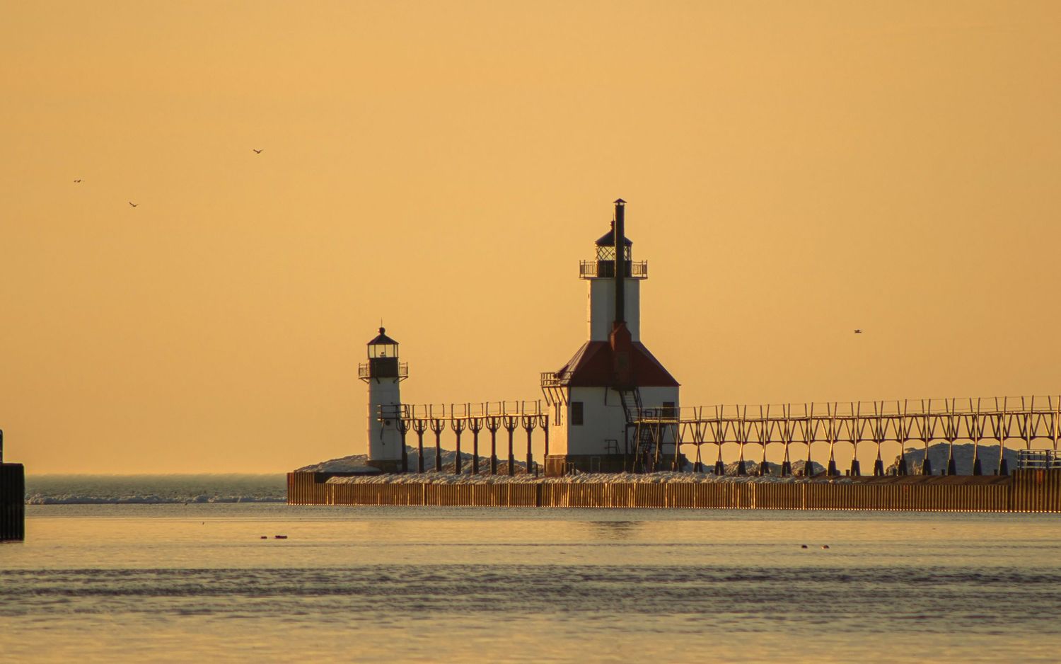St. Joseph lighthouse and pier silhouetted against golden sunset sky over calm waters.