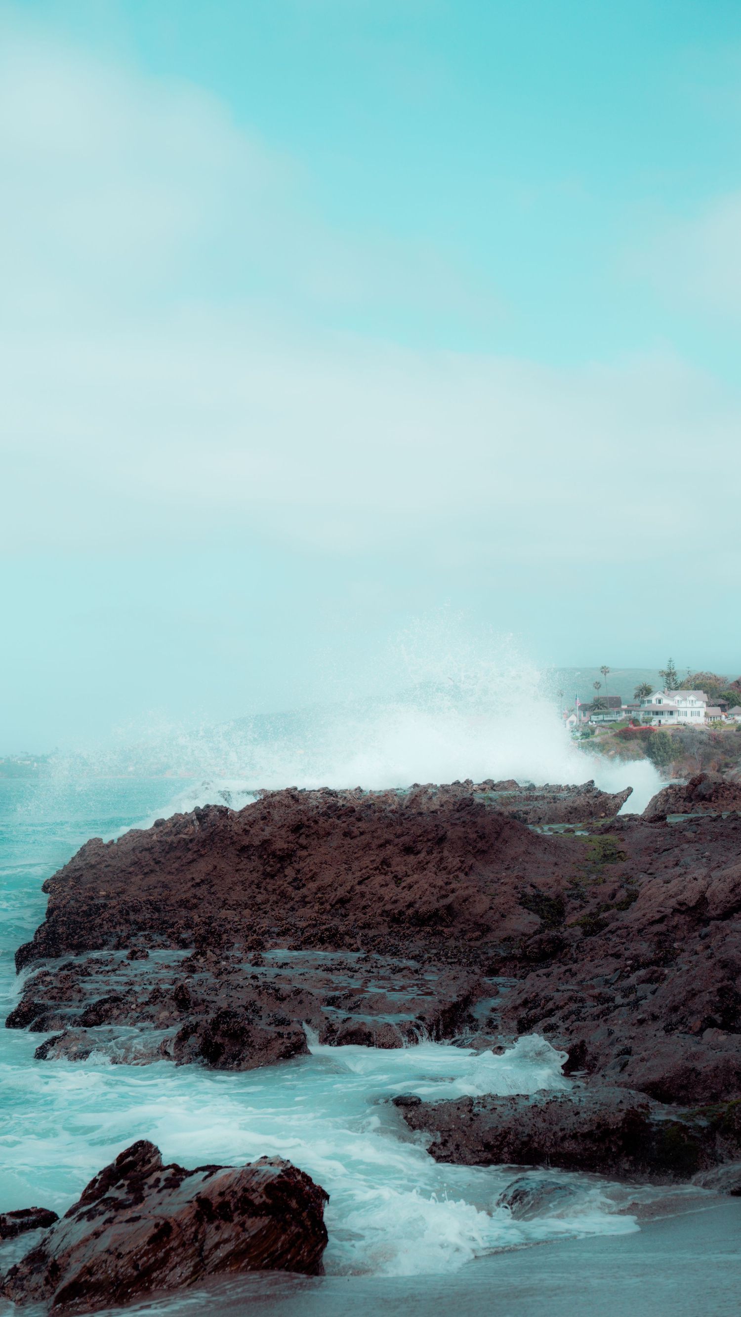 Coastal landscape showing waves crashing against volcanic rocks with white buildings visible on the cliff.