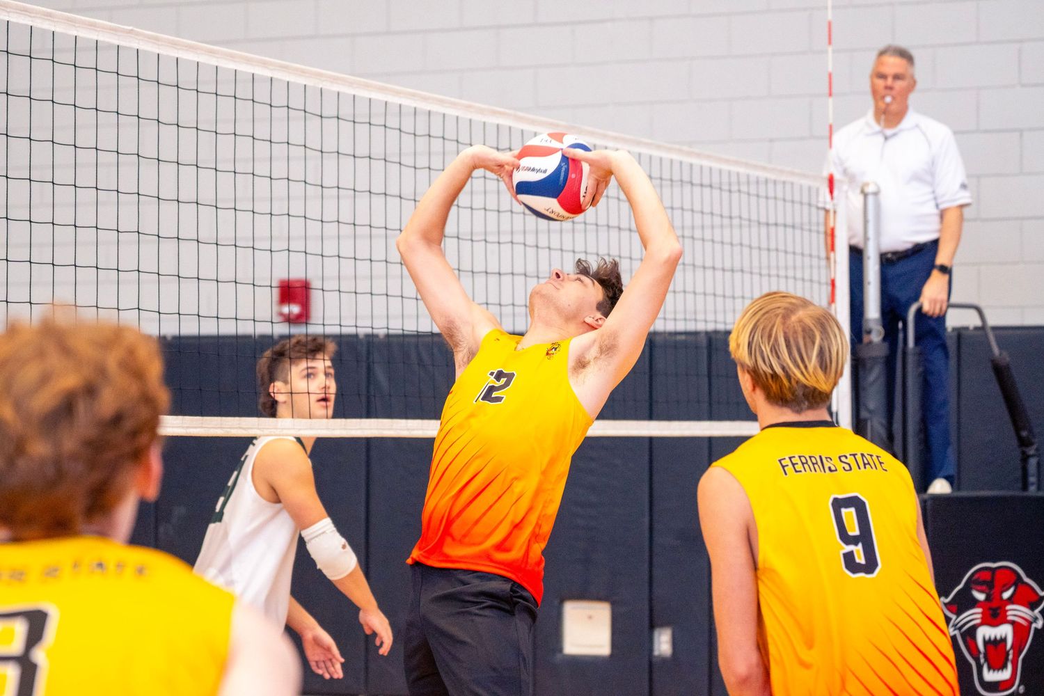 Volleyball players in yellow jerseys compete at the net during an indoor match while a referee watches from the sideline.