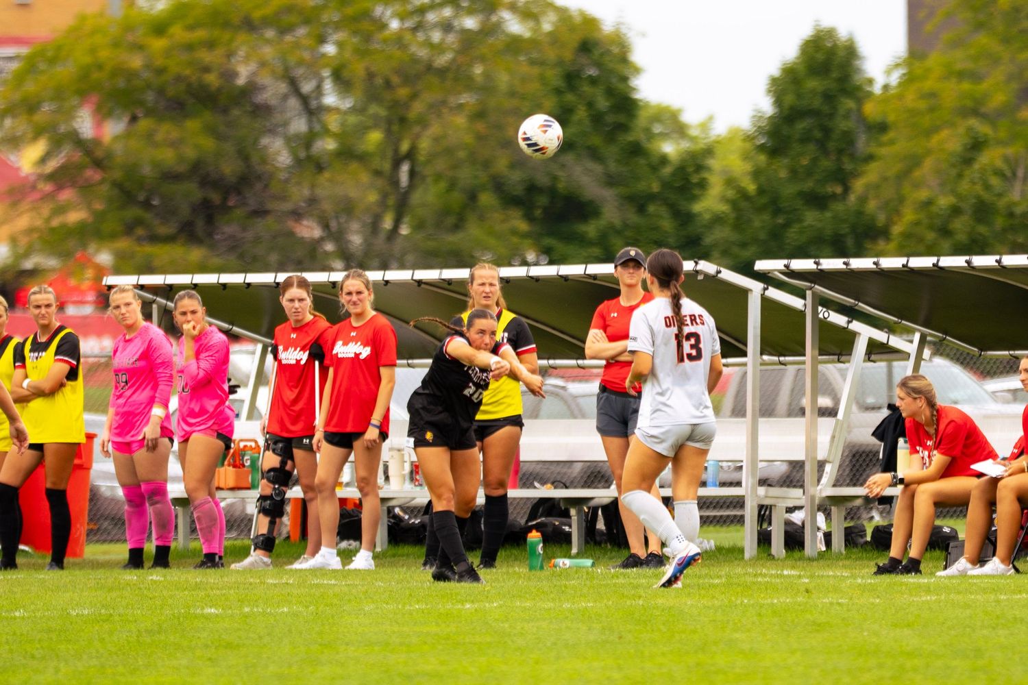 Women's soccer players in red, pink, and black uniforms watch a ball in mid-air during a match on a grass field near a sideline shelter.