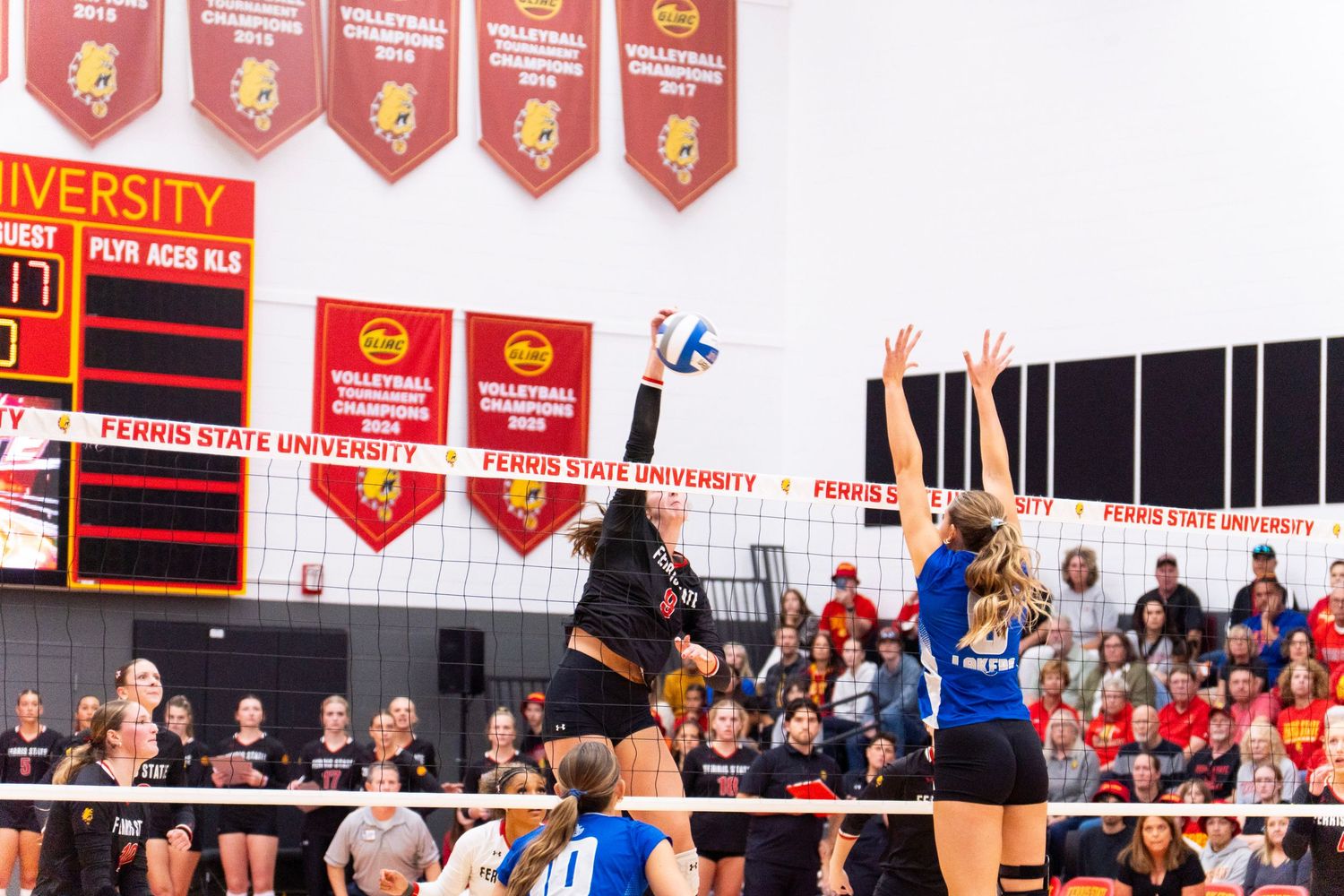 Ferris State University volleyball player in black jumps to spike the ball at the net as a blocker in blue defends before a packed crowd.