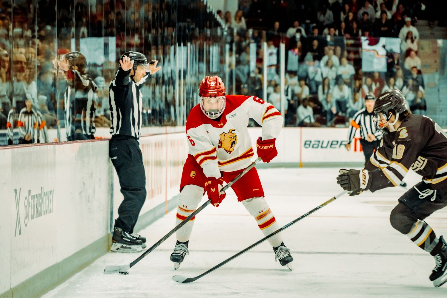 Ice hockey player in a red and white jersey skates with his stick down while an opponent in dark gear defends and a referee watches nearby.