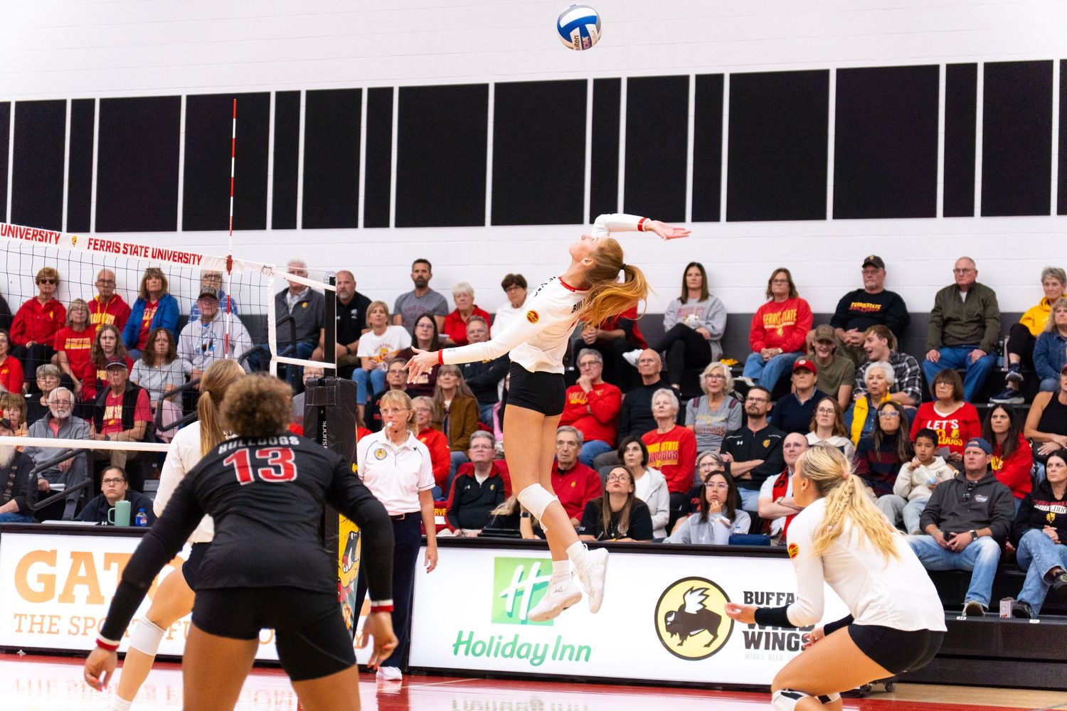 Volleyball player in white jumps to hit the ball at the net as a defender in black number 13 watches and fans in red cheer from the stands.