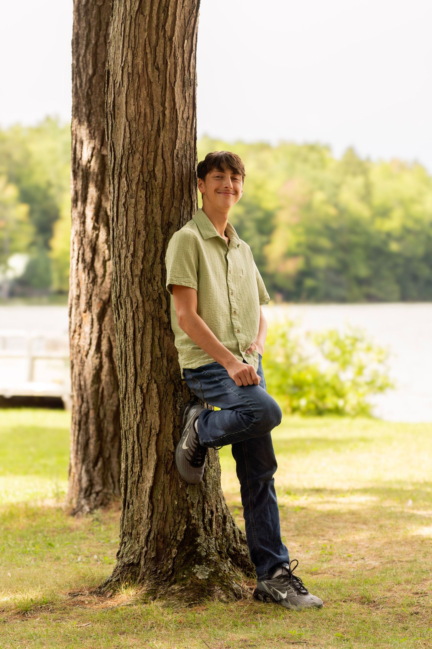 Teen in a green short-sleeve shirt and jeans leans against a large tree trunk on a grassy lawn with a calm lake in the background.