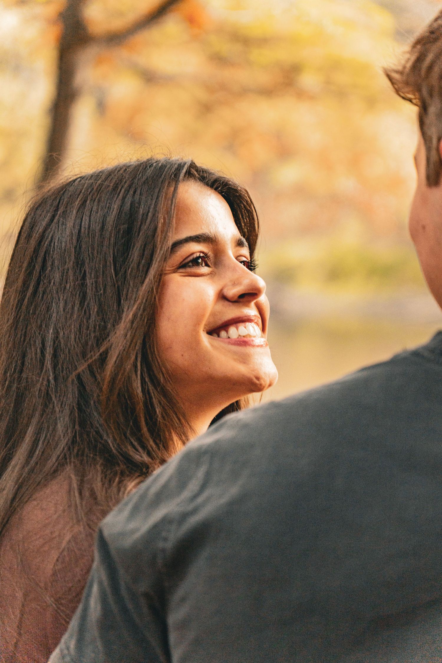 Smiling woman with long dark hair looks up at a man in the foreground against a blurred autumn park background.
