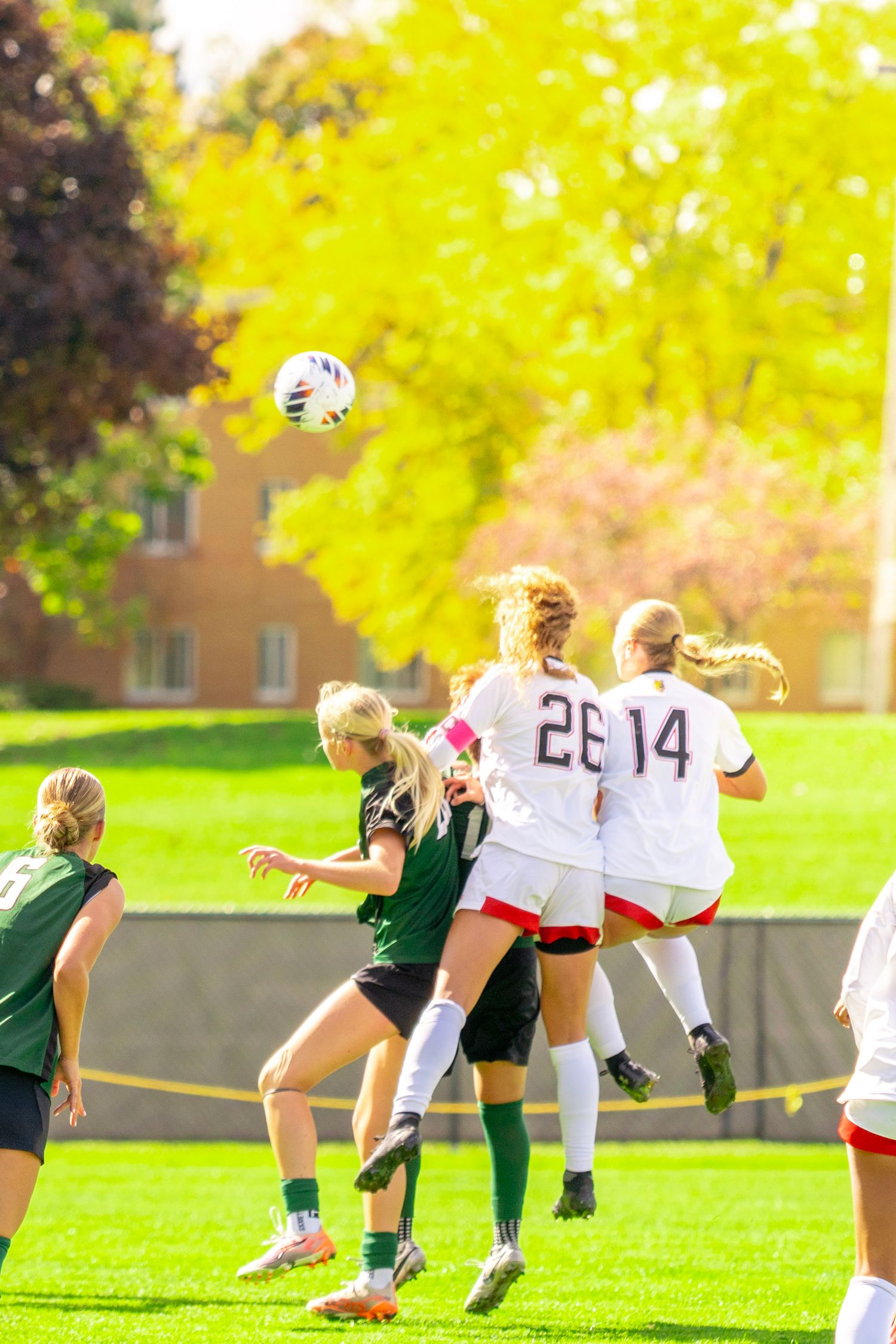 Women's college soccer players in white and green uniforms leap for a header as the ball flies above the field on a sunny autumn day.