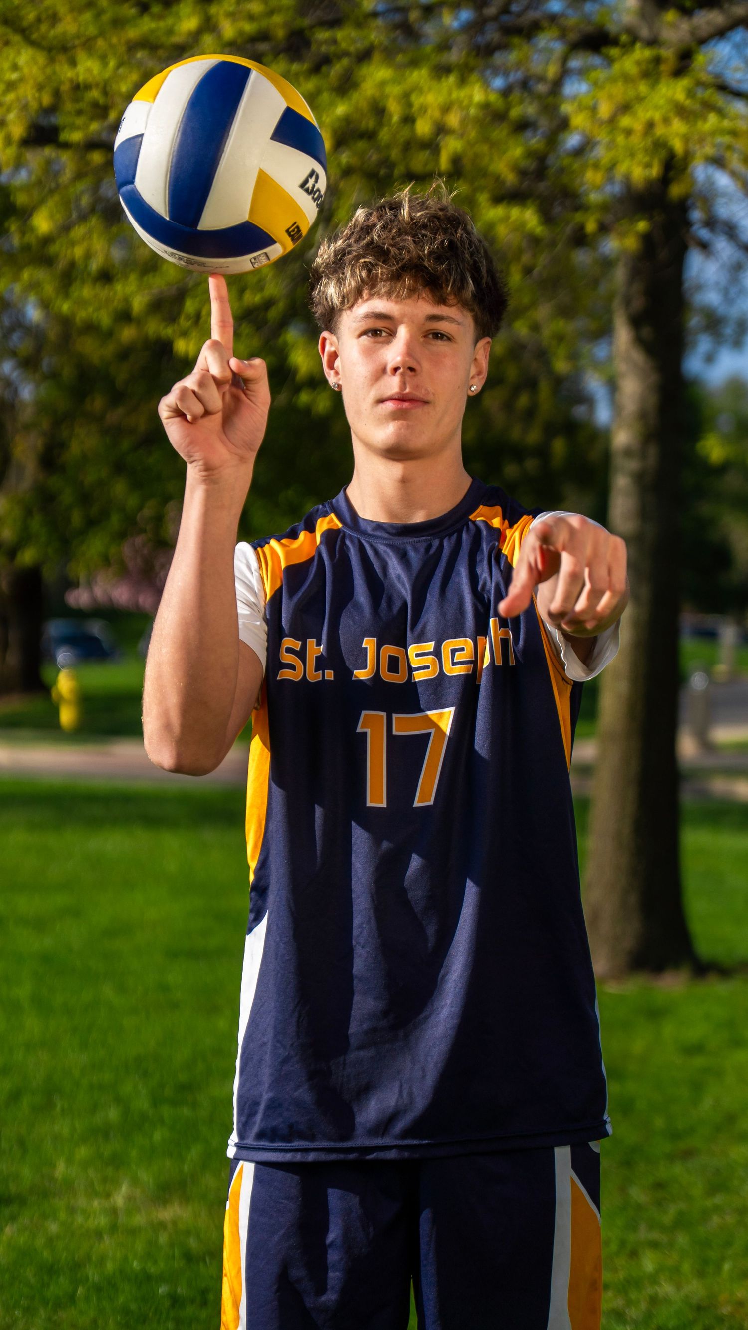 St. Joseph player number 17 balances volleyball on finger while pointing at camera outdoors.