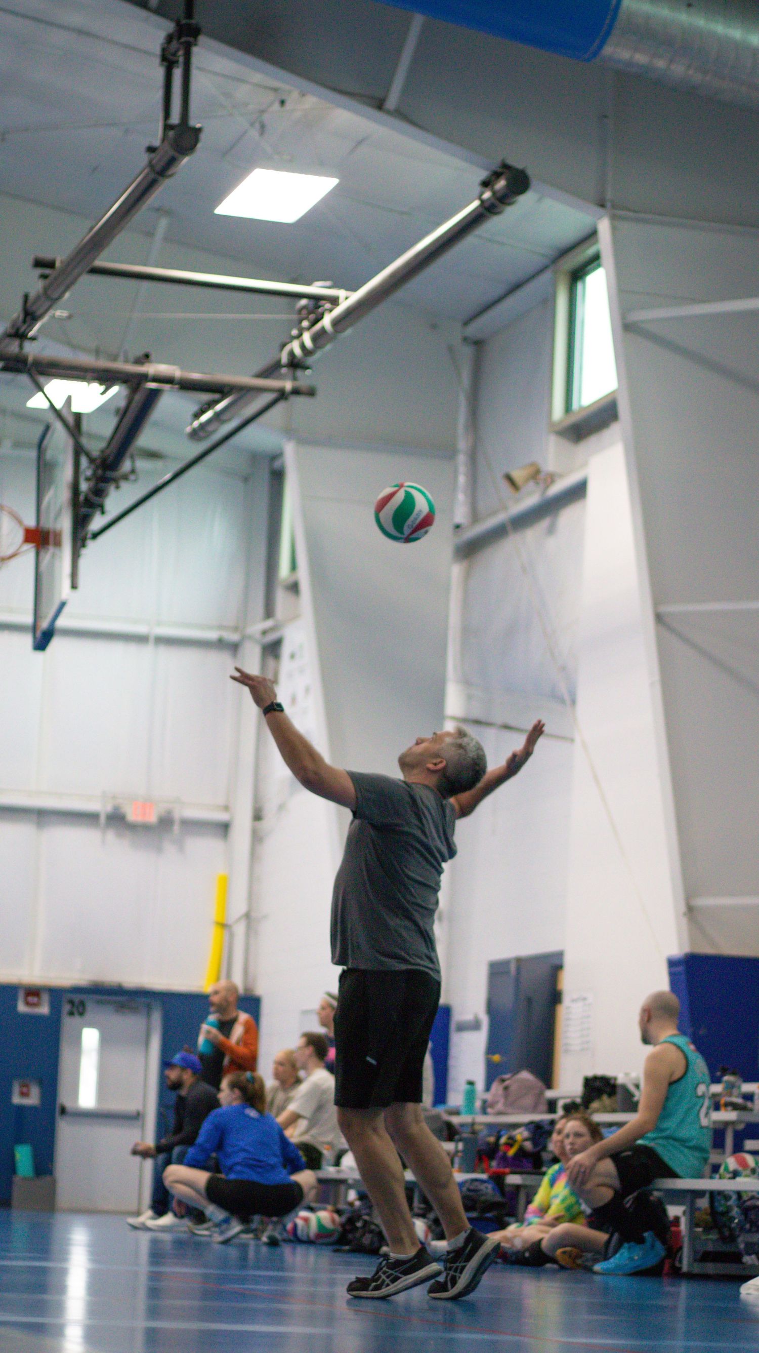 Player jumps to serve volleyball in bright indoor gymnasium with spectators watching.