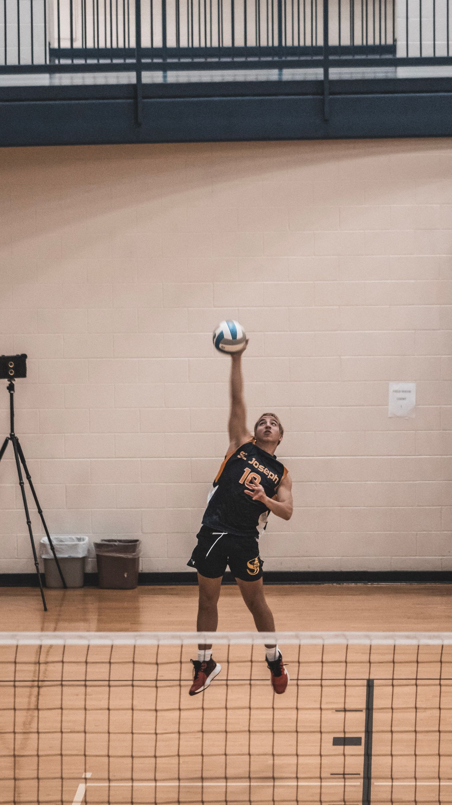 Player jumps to serve volleyball in gymnasium during match, with net and wall visible.