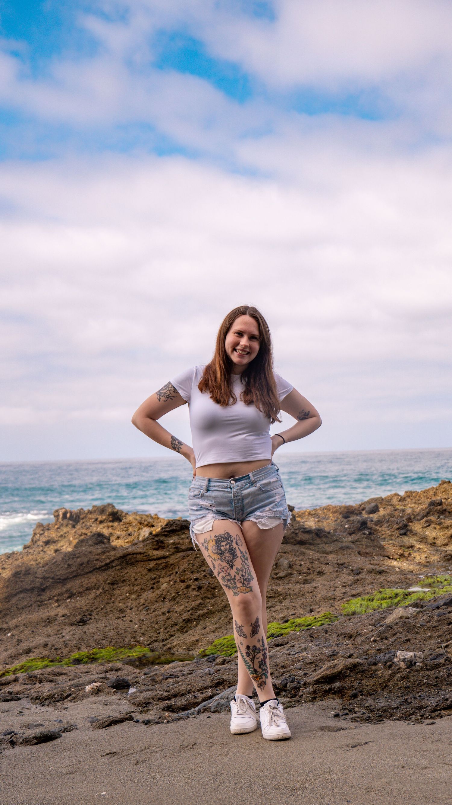 Person posing on rocky coastline with ocean waves and blue sky in the background.