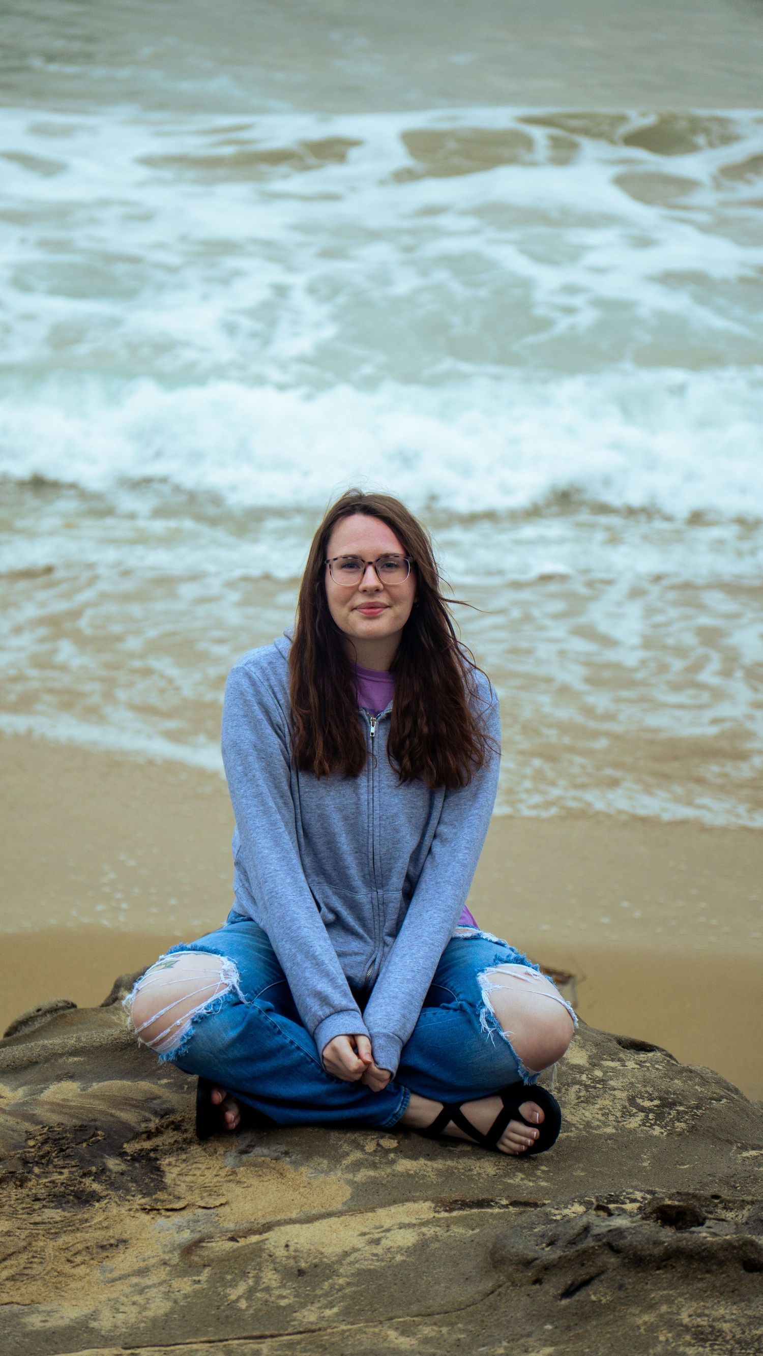 Someone sits on coastal rocks wearing denim and a gray hoodie as waves crash behind them.