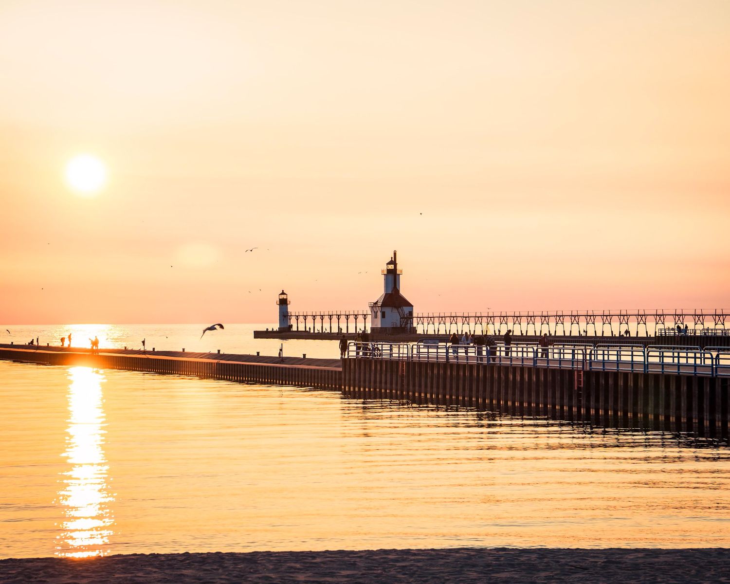 Long pier stretches into calm waters during golden sunset with lighthouse at the end.