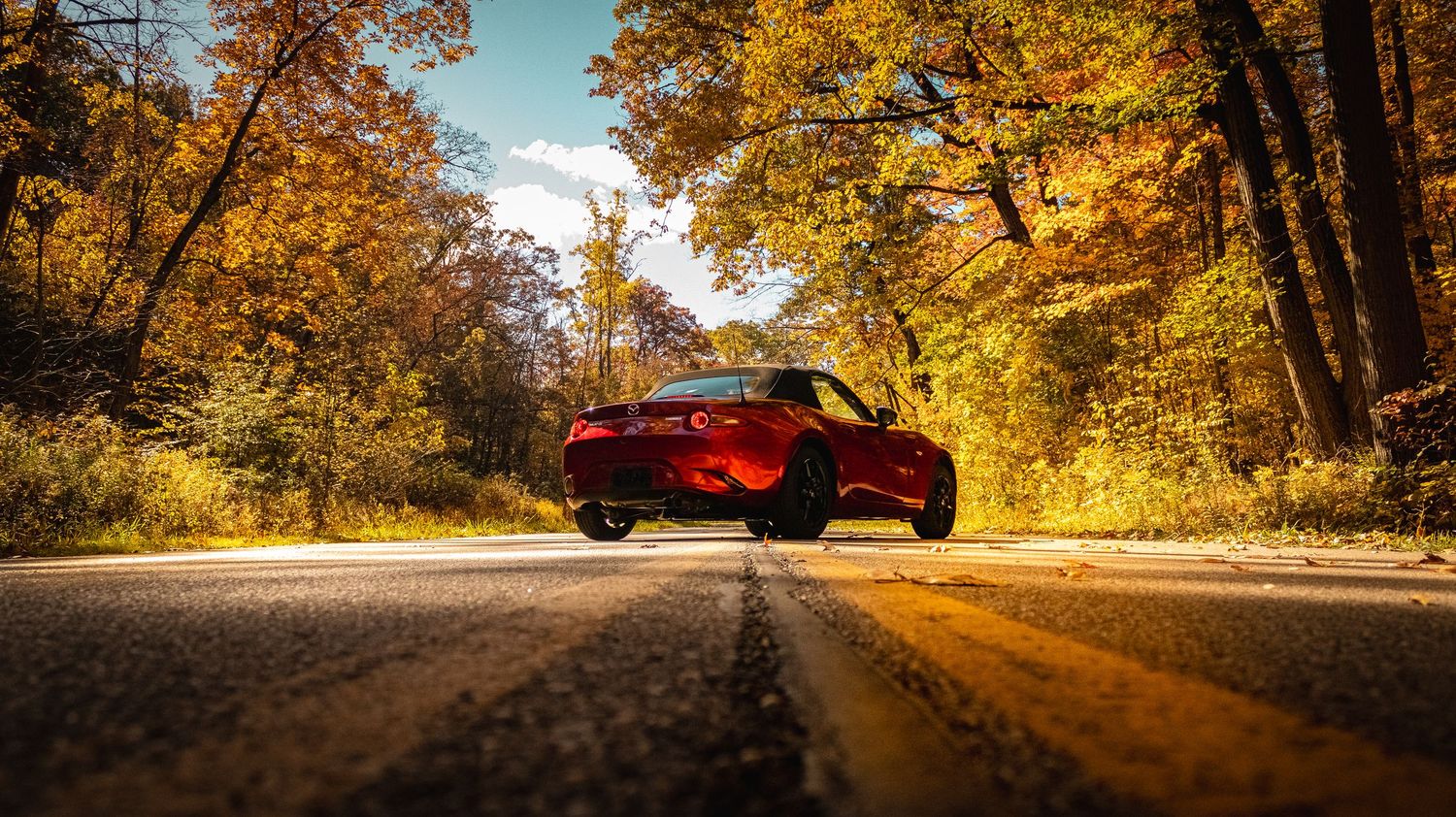 Red Mazda Miata on country road framed by vibrant fall foliage and sunlight.