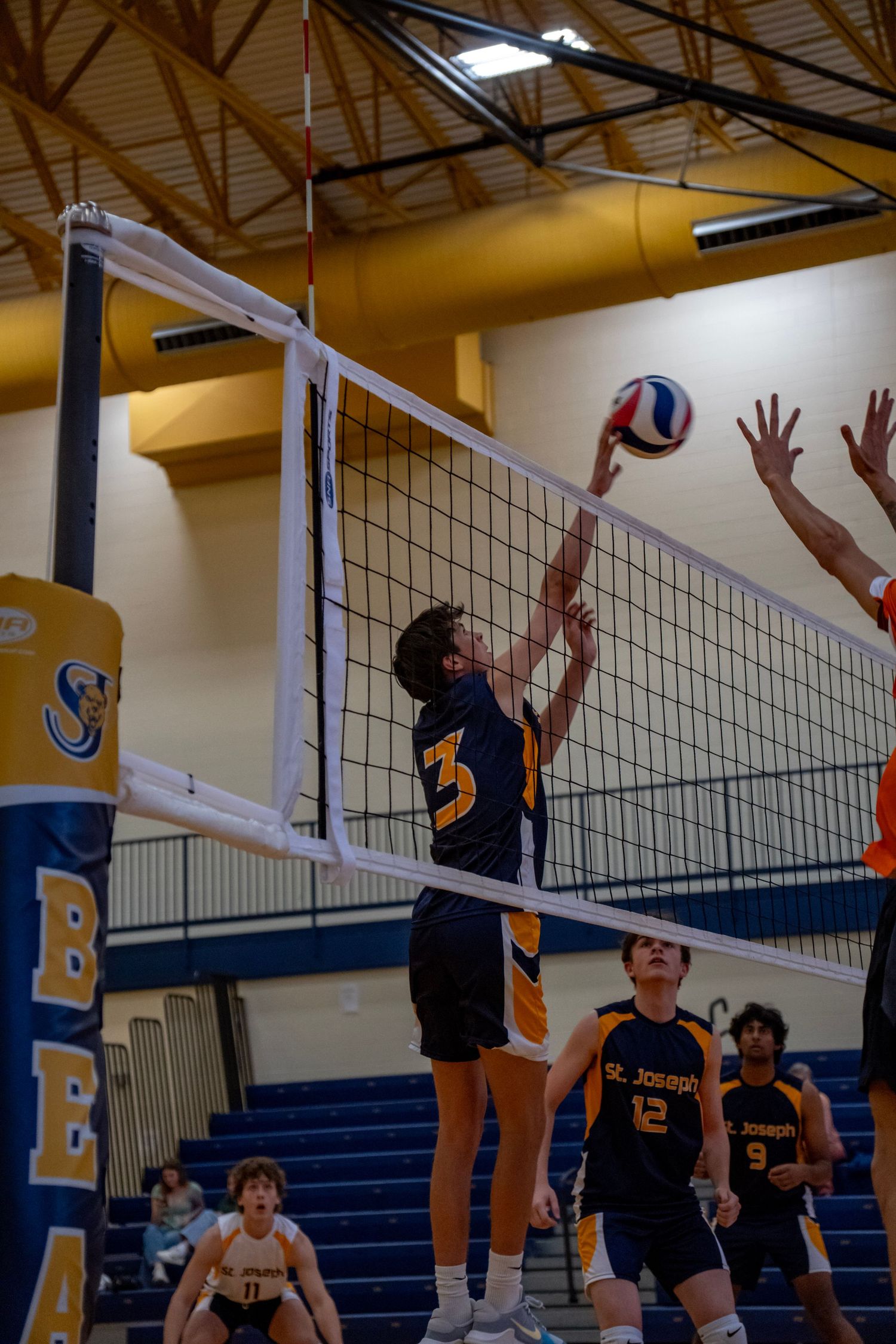 Volleyball player in navy jersey reaches up to spike the ball over the net during indoor game.
