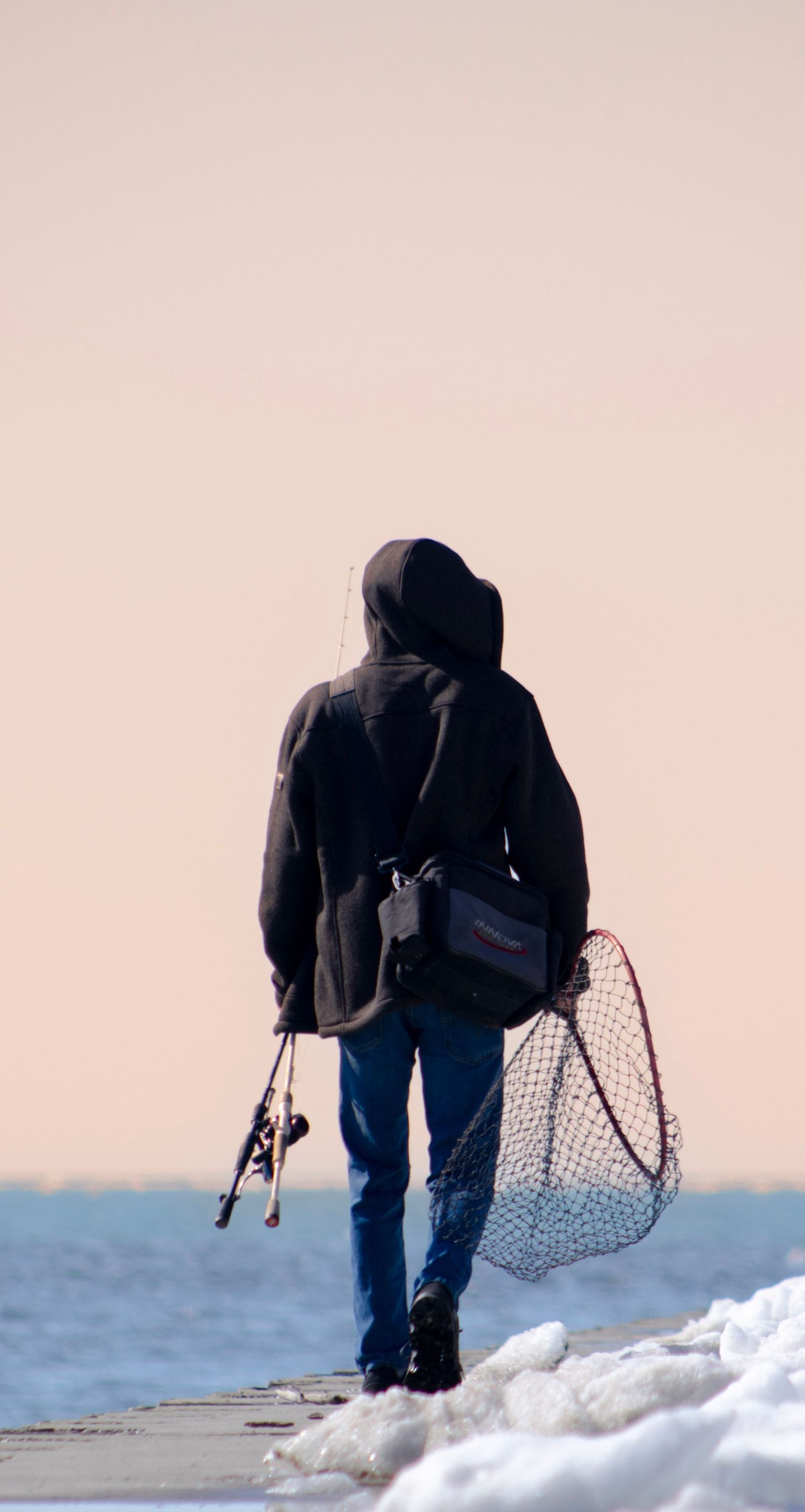 Figure with fishing gear walking on snowy pier against pink sky.