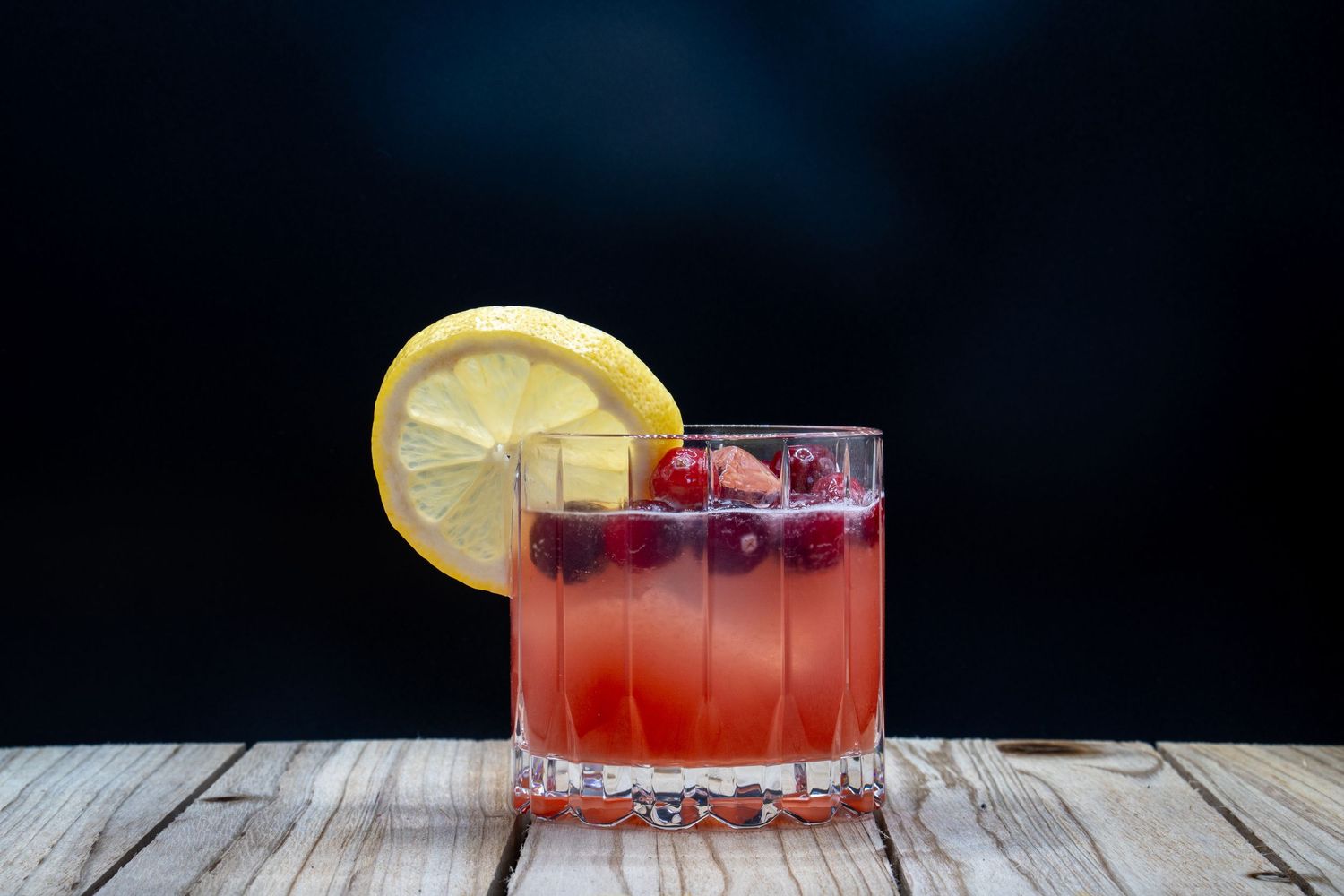 Red cocktail garnished with lemon slice and berries in a rocks glass on wooden surface.