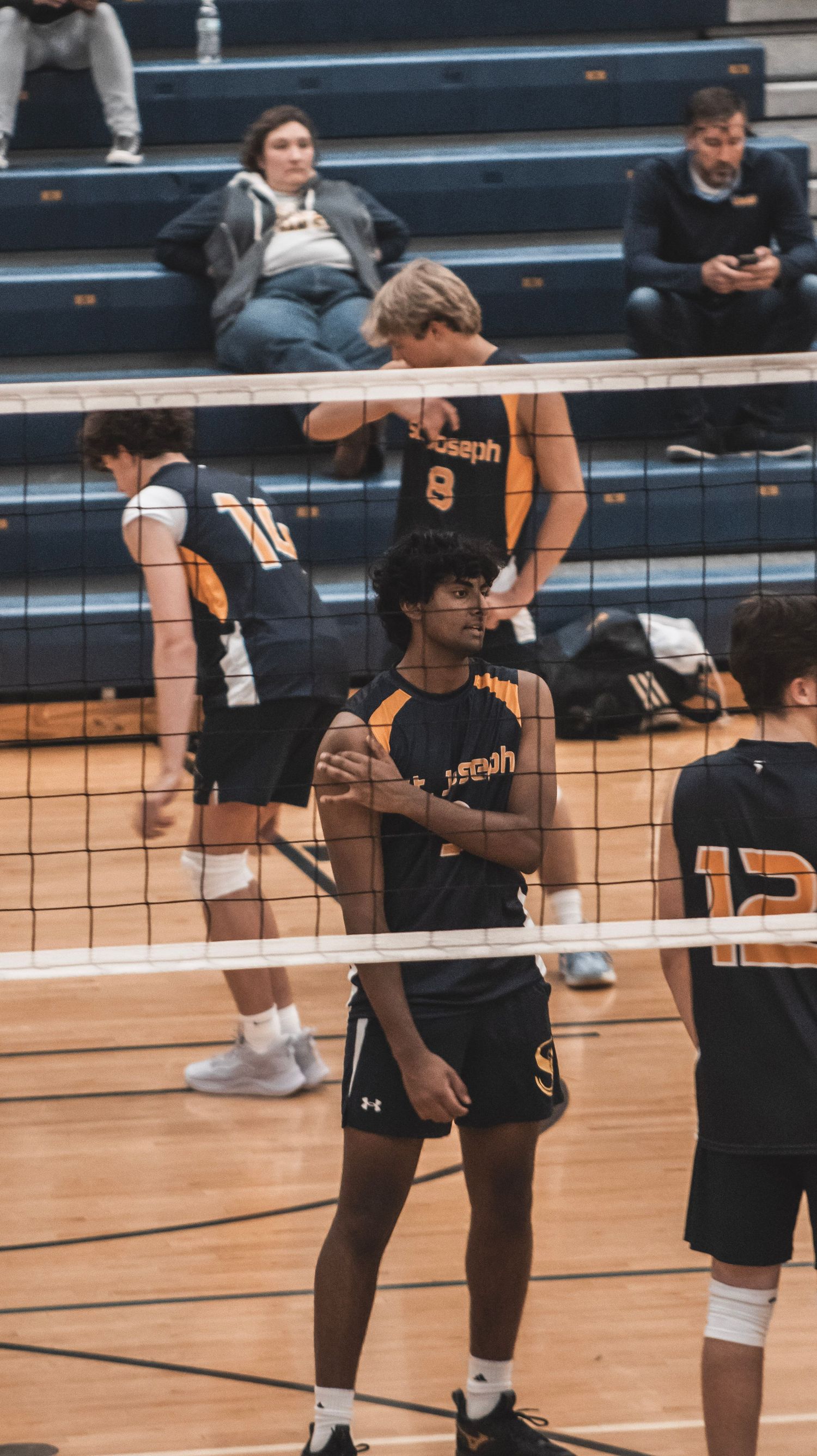 Players in black and orange uniforms standing near the volleyball net during a break in play.