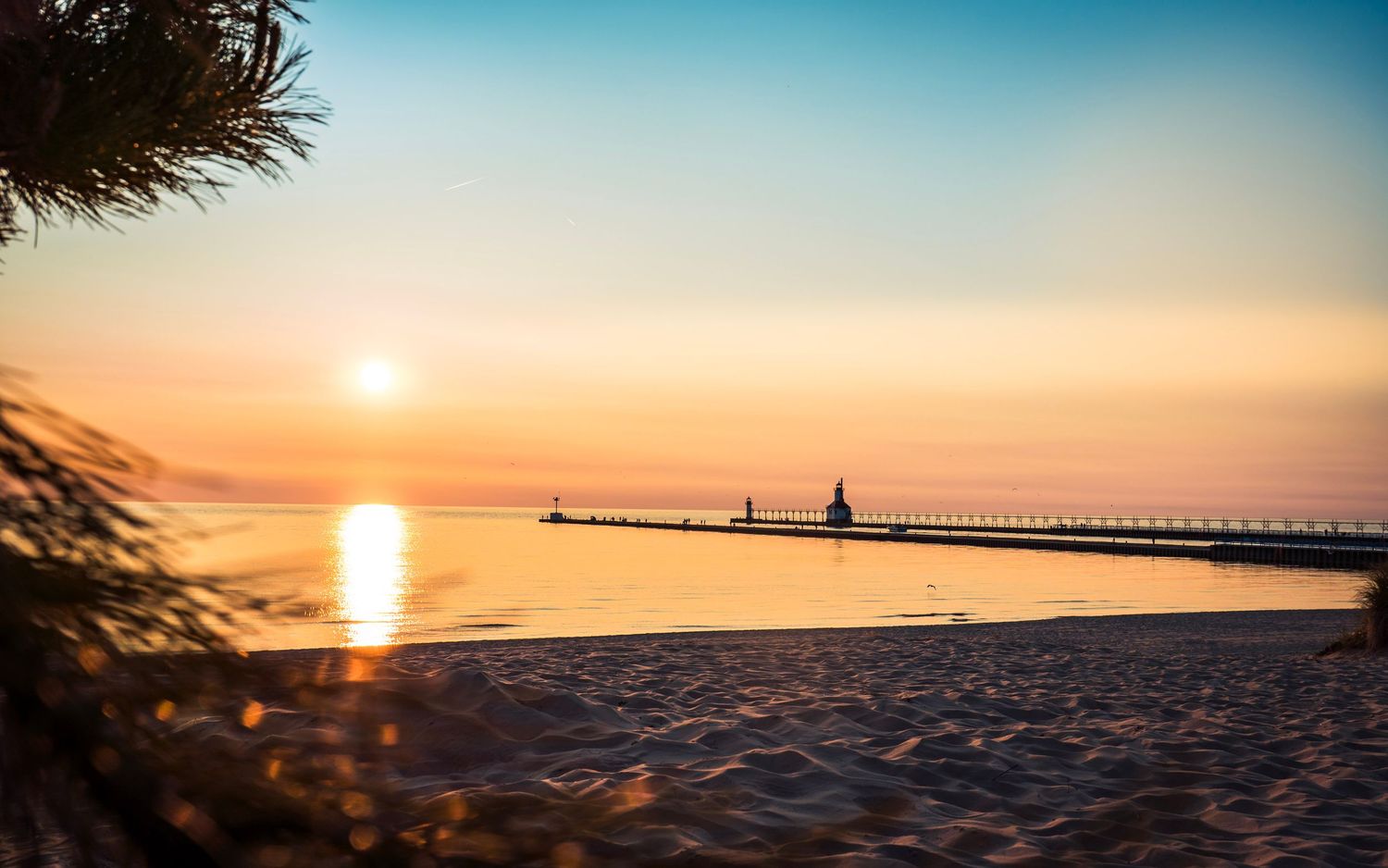 Michigan lighthouse and pier reflecting golden sunlight during sunset over Lake Michigan.