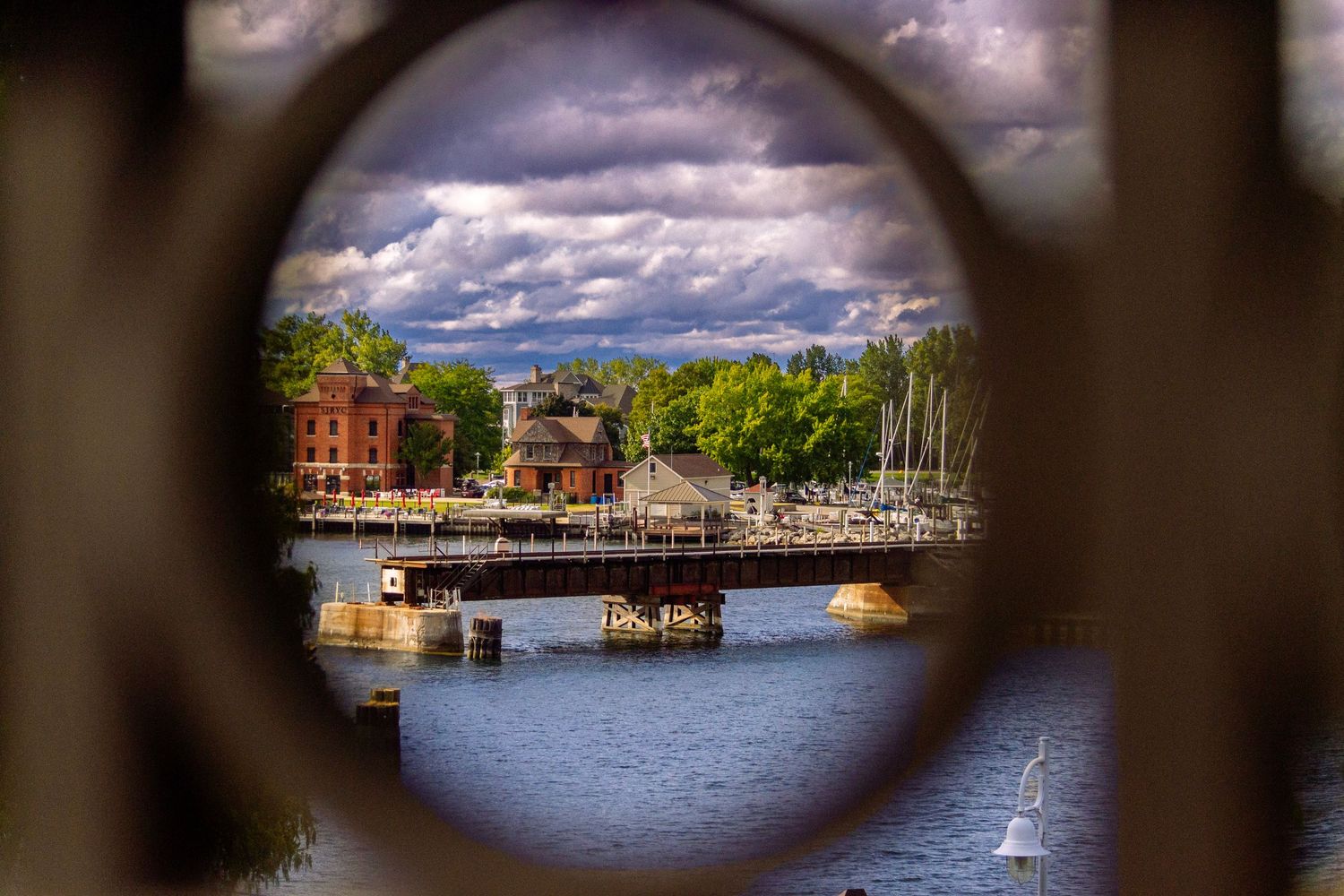 View through a circular frame of a harbor with boats and buildings under dramatic cloudy skies.
