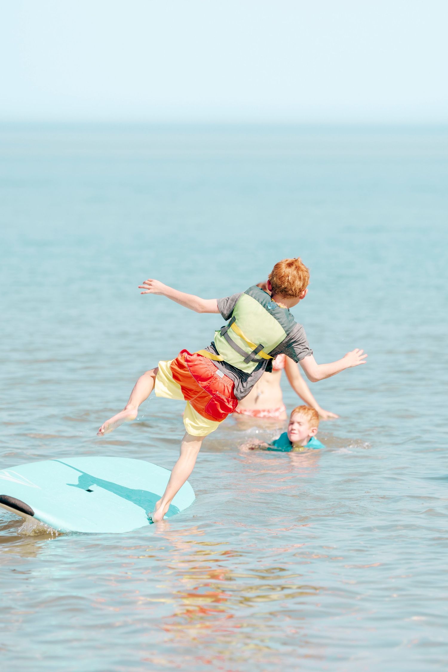 A person wearing a life vest attempts to balance while learning to skim board in calm ocean waters.
