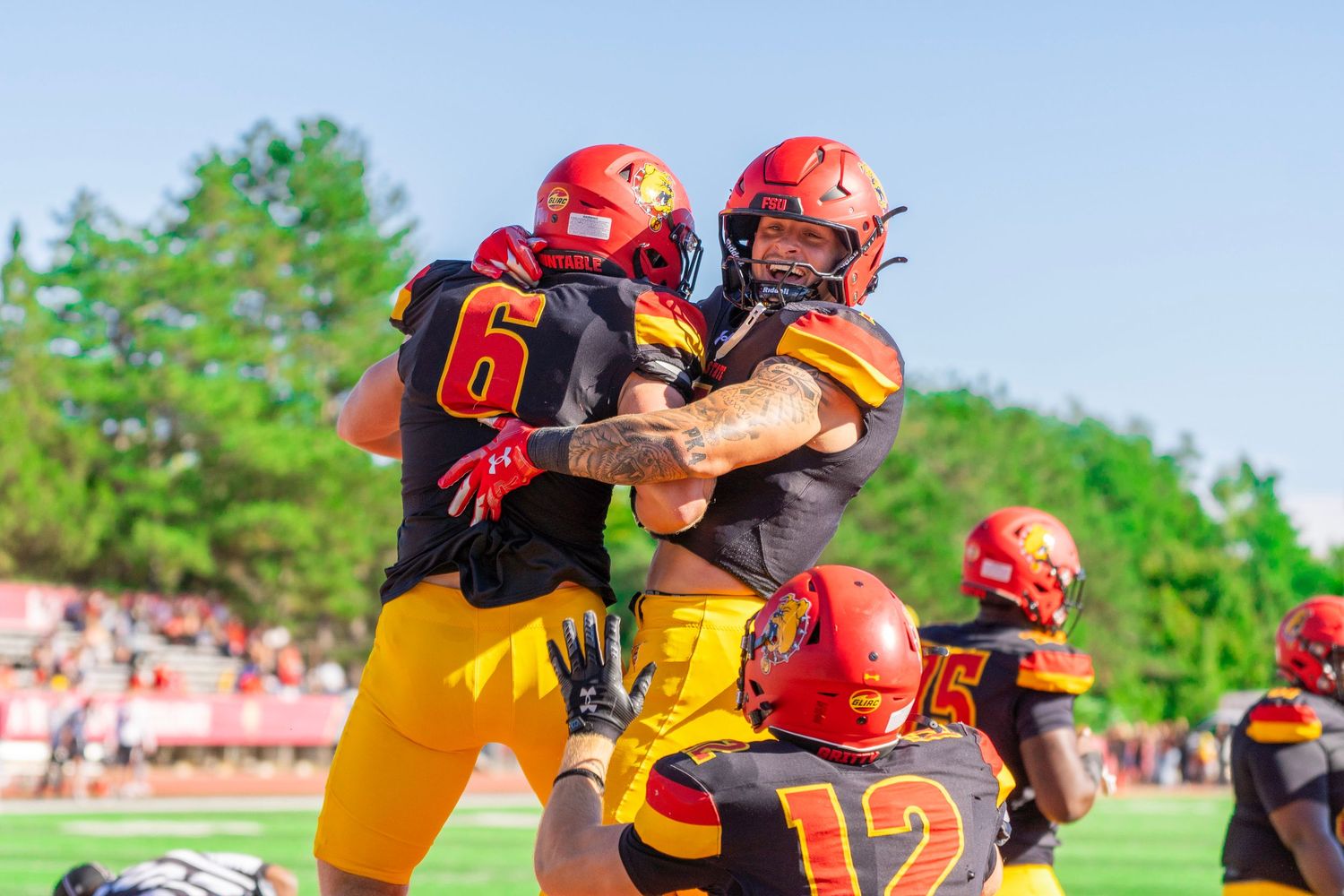Football players in red helmets and black and gold uniforms celebrate together on the field during a game on a sunny day.