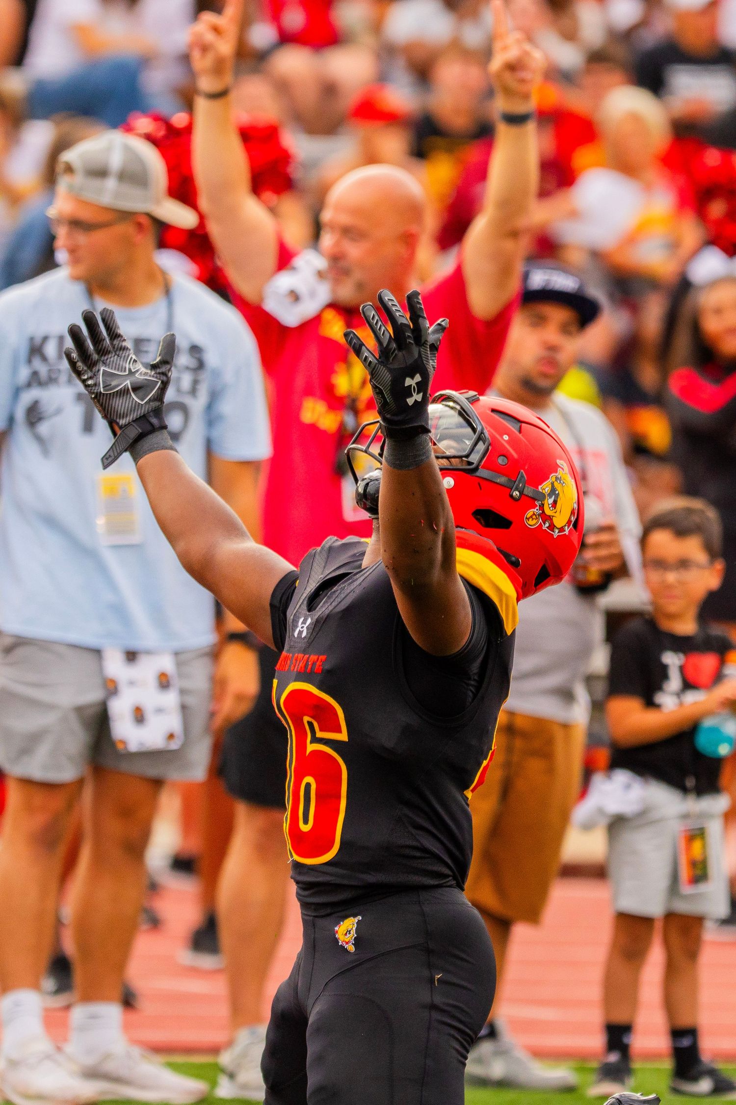 Football player in a black number 6 jersey and red helmet raises his gloved hands in celebration as fans cheer behind him.