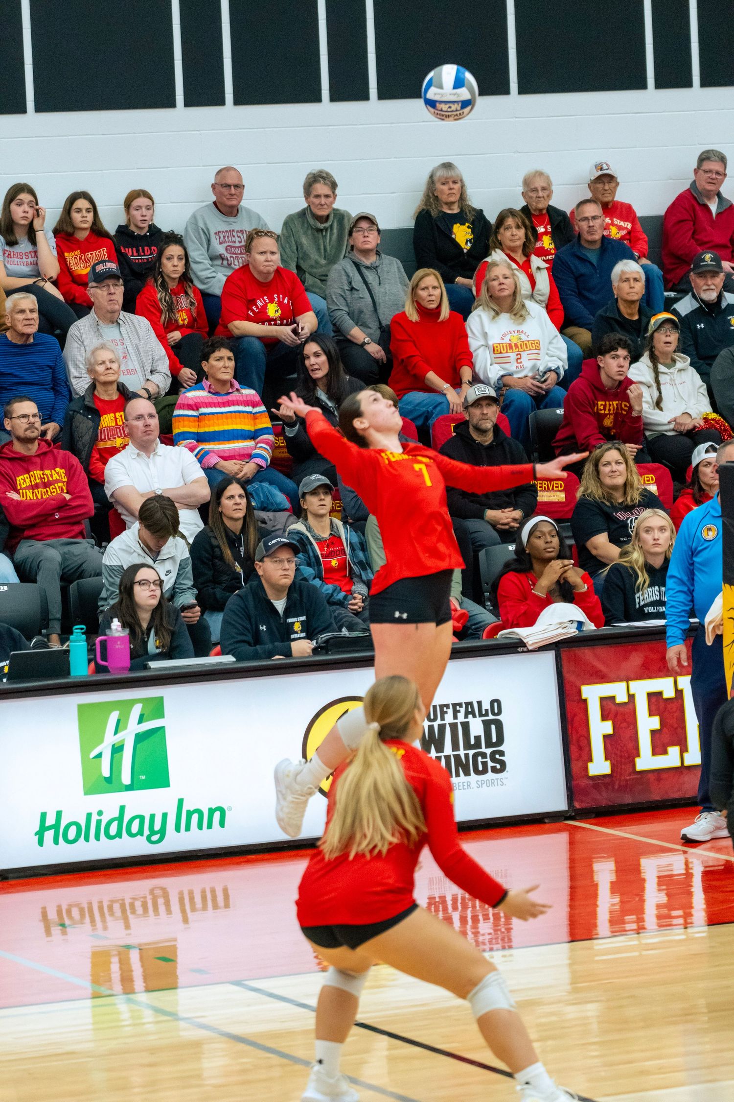 College volleyball players in red uniforms compete during a match with spectators watching from the bleachers.