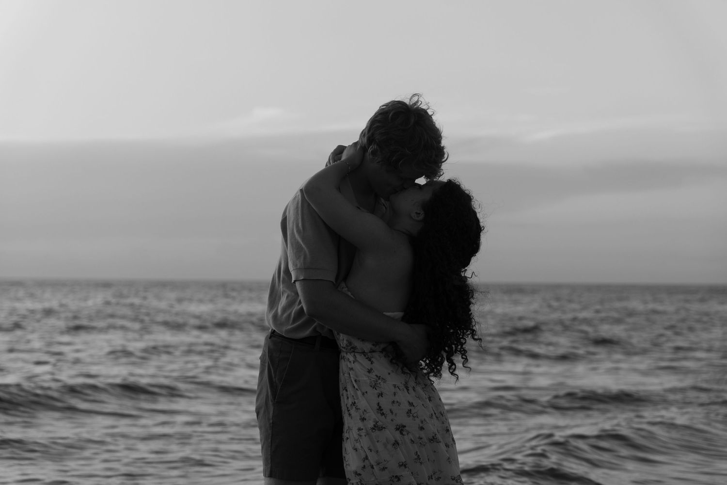 A romantic couple embraces on the beach at sunset in a black and white silhouette against the ocean.