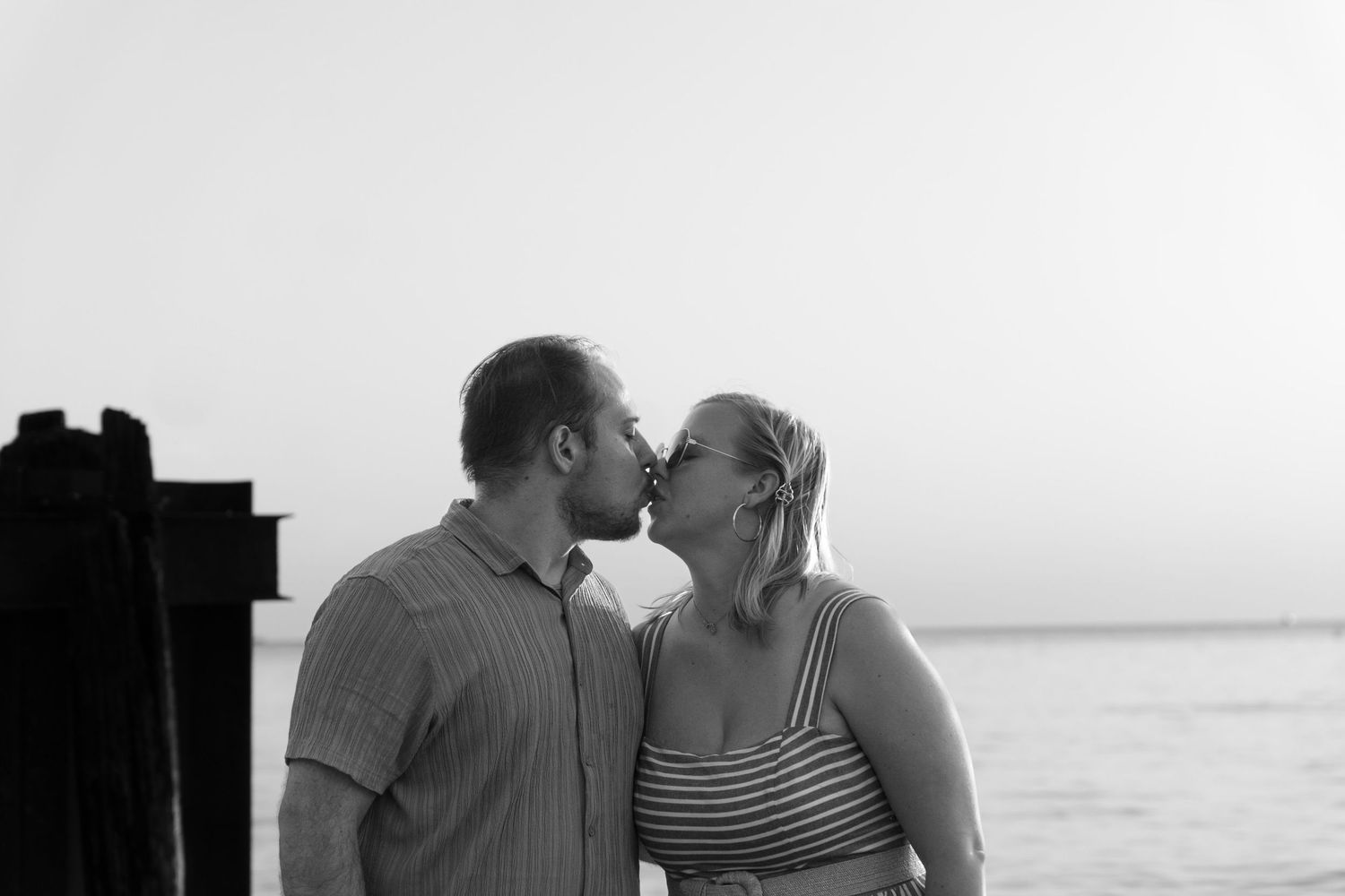 A couple shares a tender kiss while standing near the water in black and white.