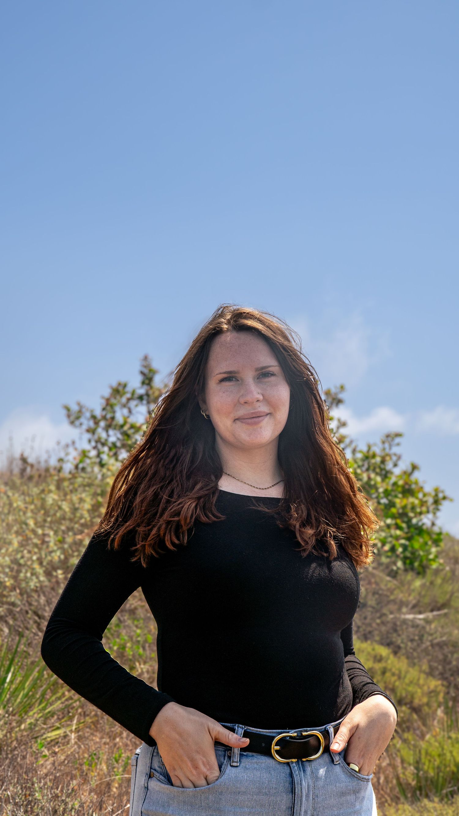 Person in black top and jeans posing outdoors against blue sky and green foliage.